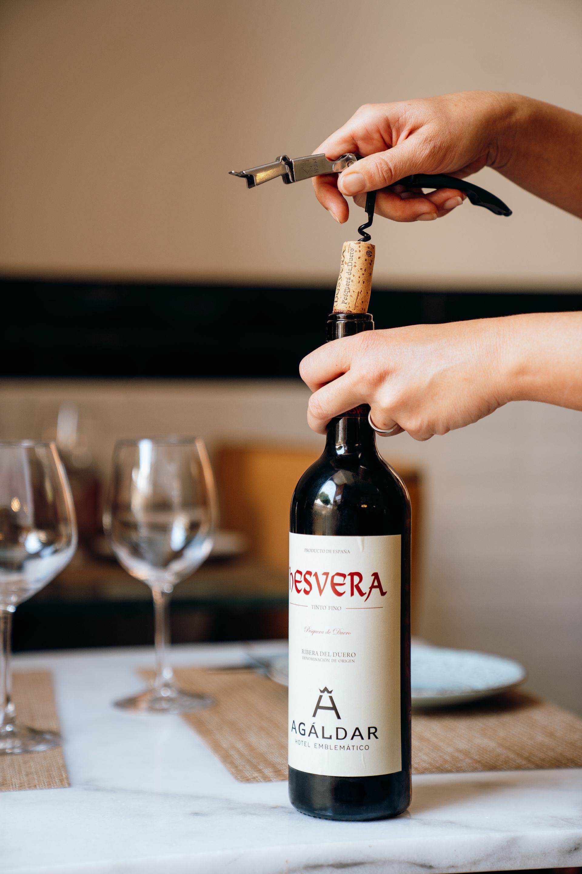 Unas manos descorchan una botella de vino en una mesa, con vasos vacíos y botellas de vino al fondo.