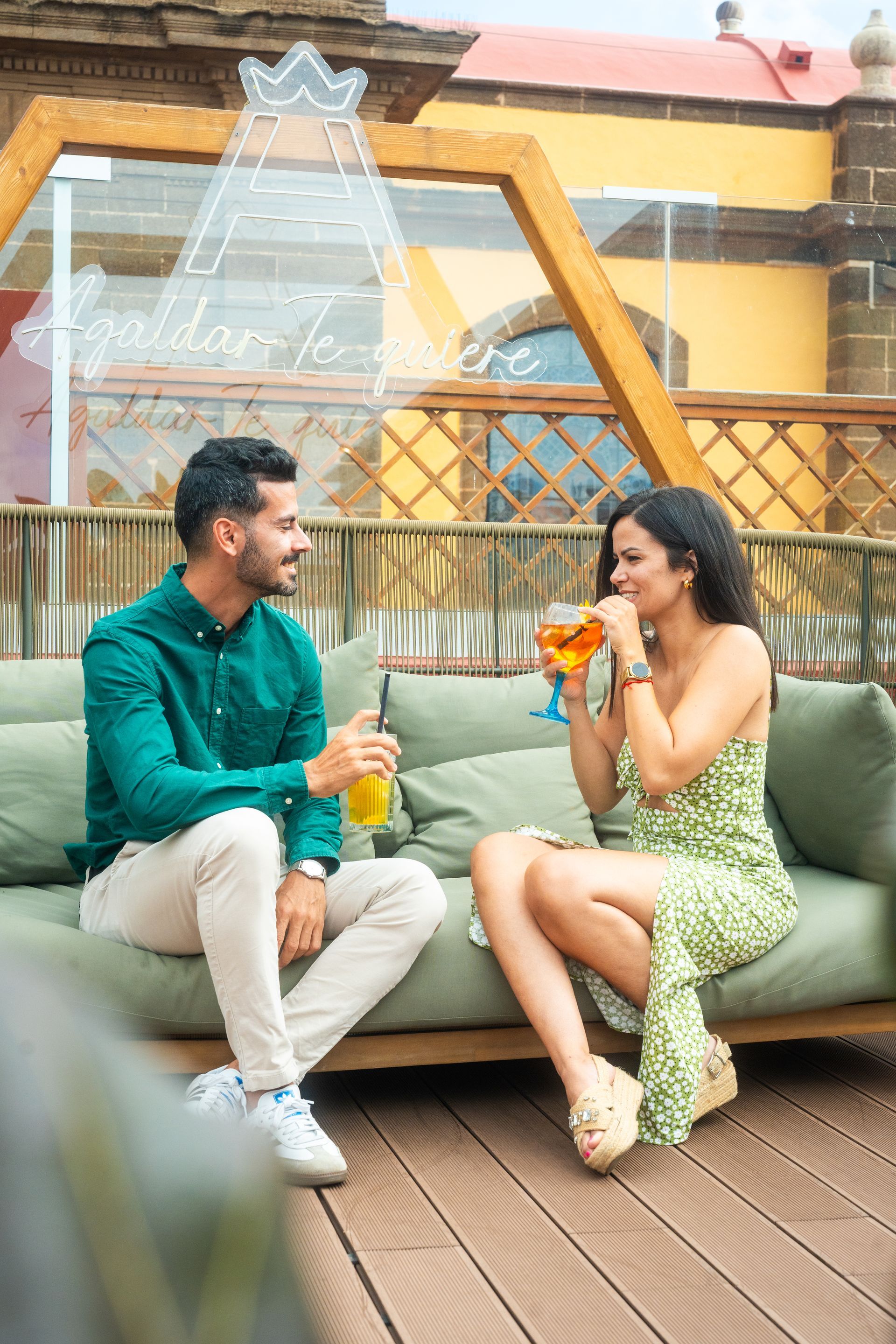 Dos personas están sentadas en un sofá de patio, sonriendo y bebiendo cócteles frente a una valla de madera.