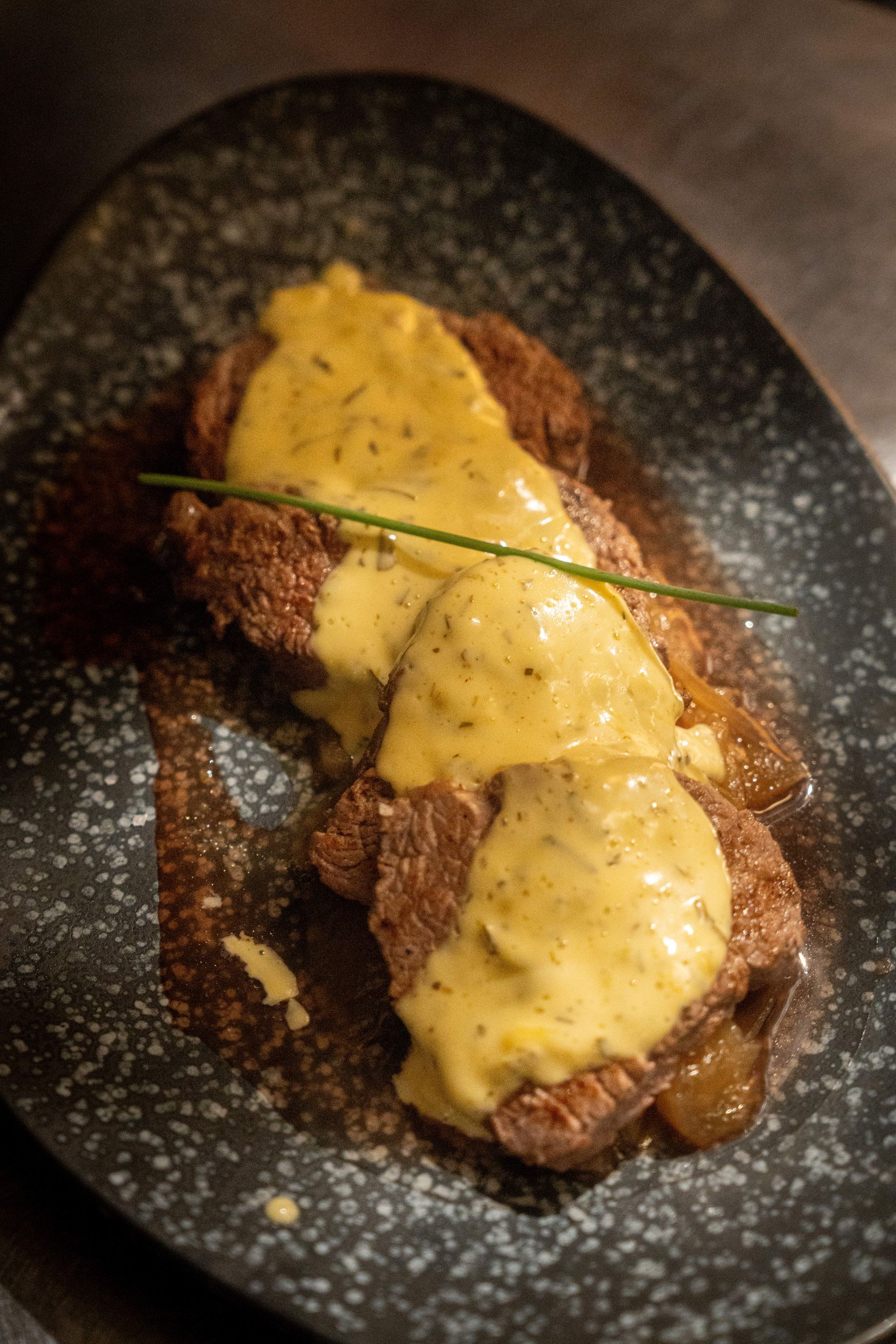 Carne a la parrilla cubierta con salsa cremosa en un plato oscuro moteado, adornada con una ramita de hierbas verdes.