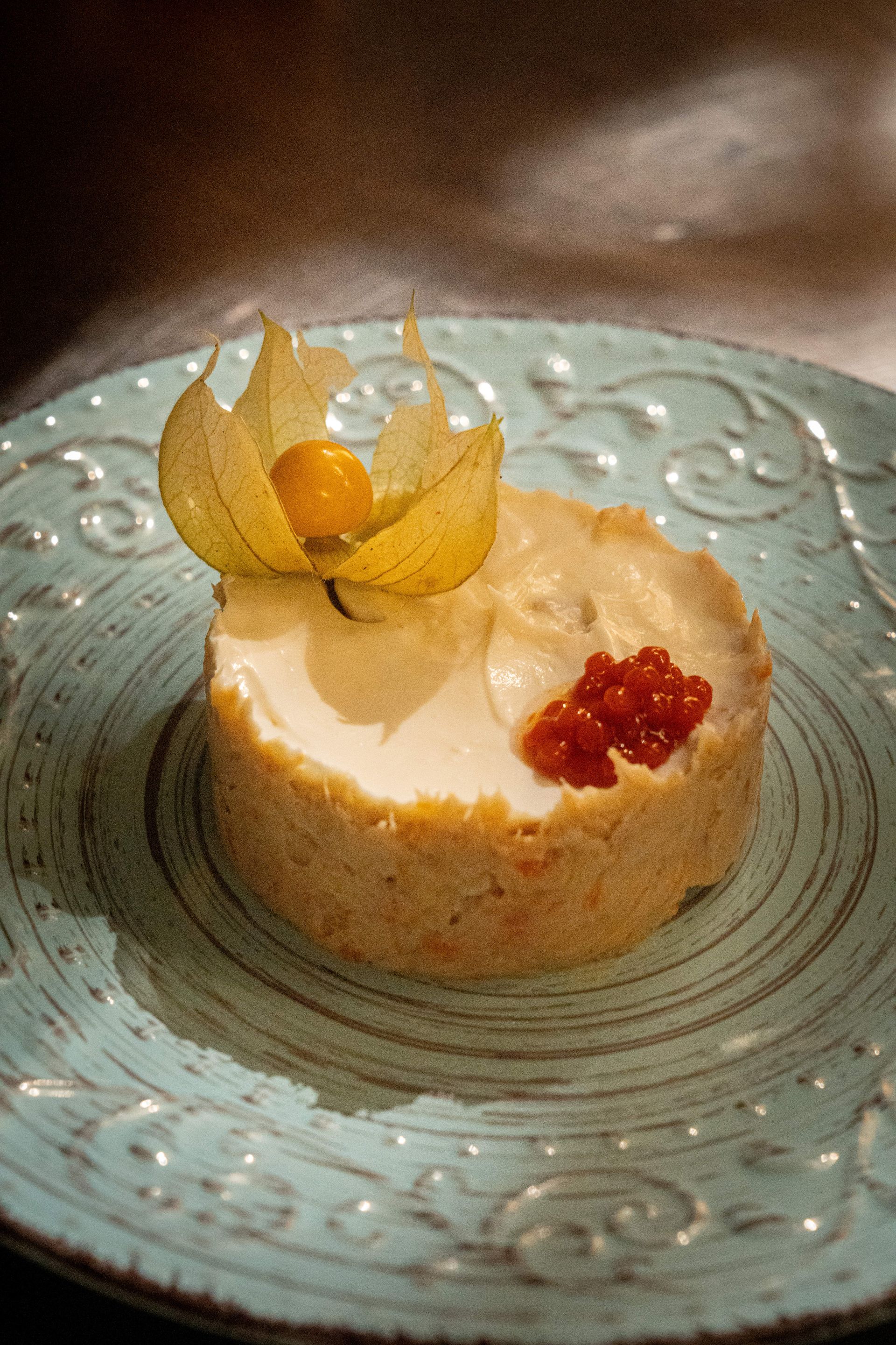 Elegante postre emplatado con crema, guarnición de frutos rojos y flor comestible amarilla sobre un plato azul verdoso.