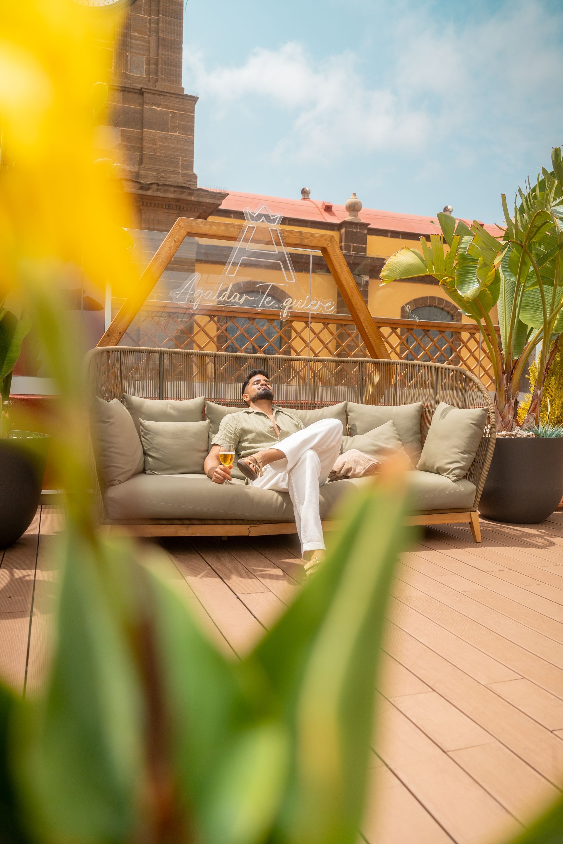 Una persona descansa en un sofá gris al aire libre, en una soleada terraza de madera, con una fuente y flores cerca.