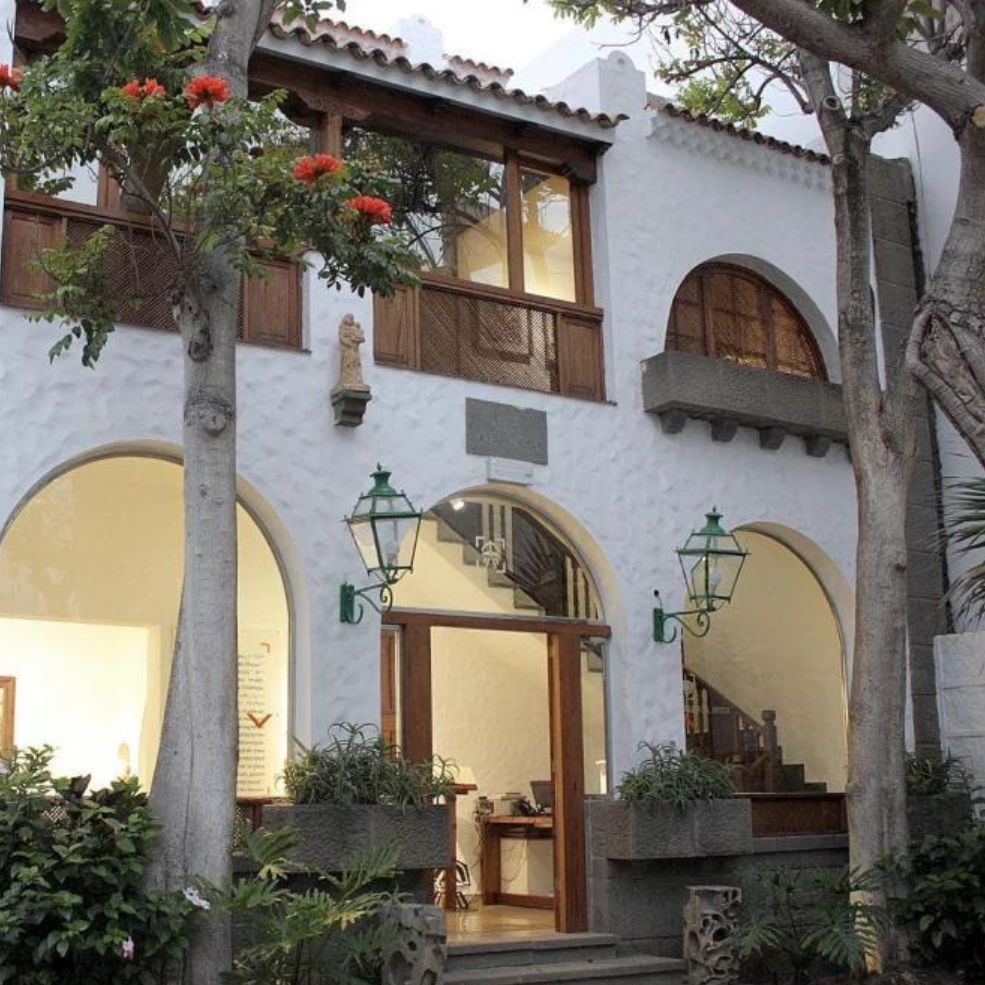 Edificio con patio de estuco blanco, arcos, faroles y árboles altos al anochecer.