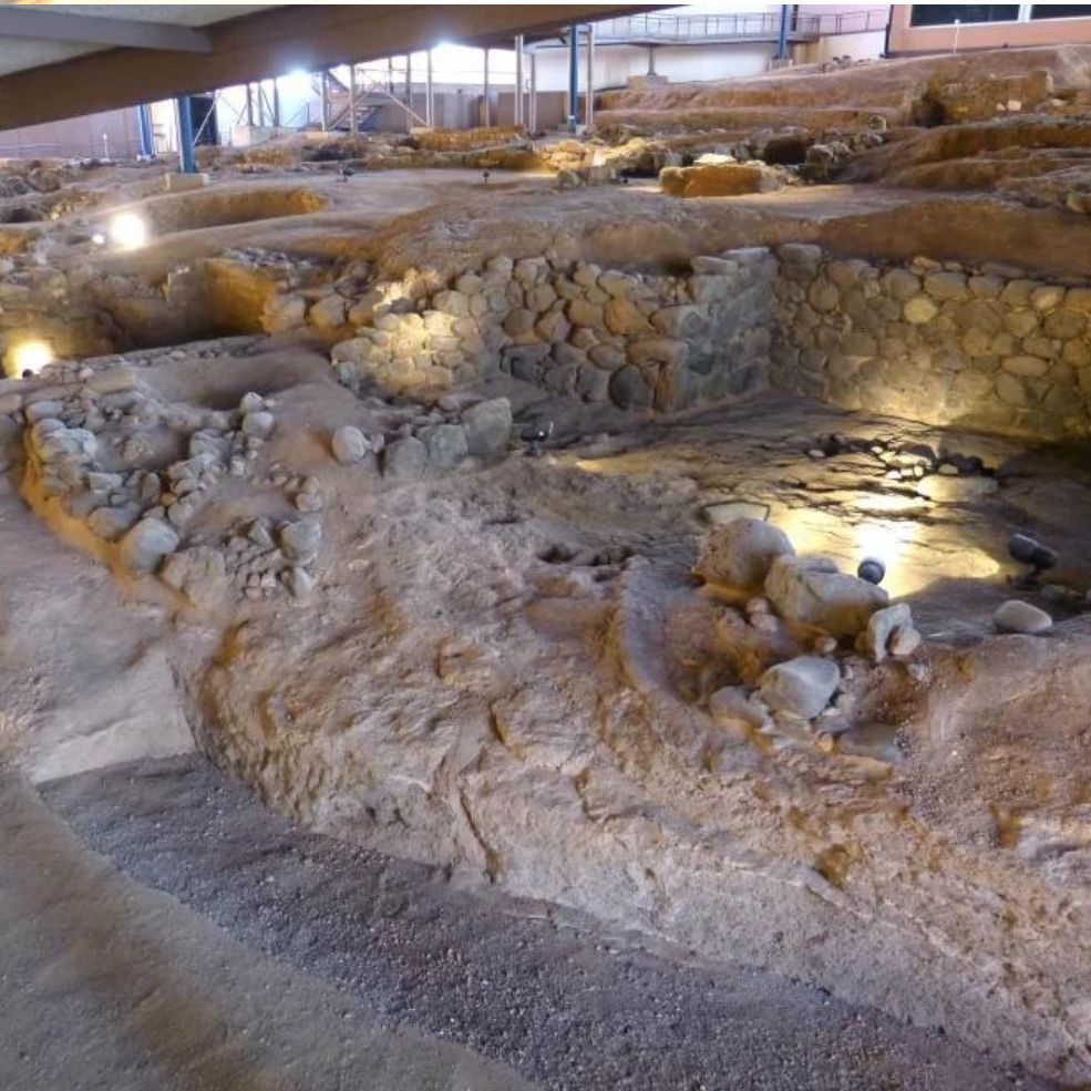 Sitio de excavación arqueológica con muros de piedra y trincheras iluminadas bajo un cobertizo.