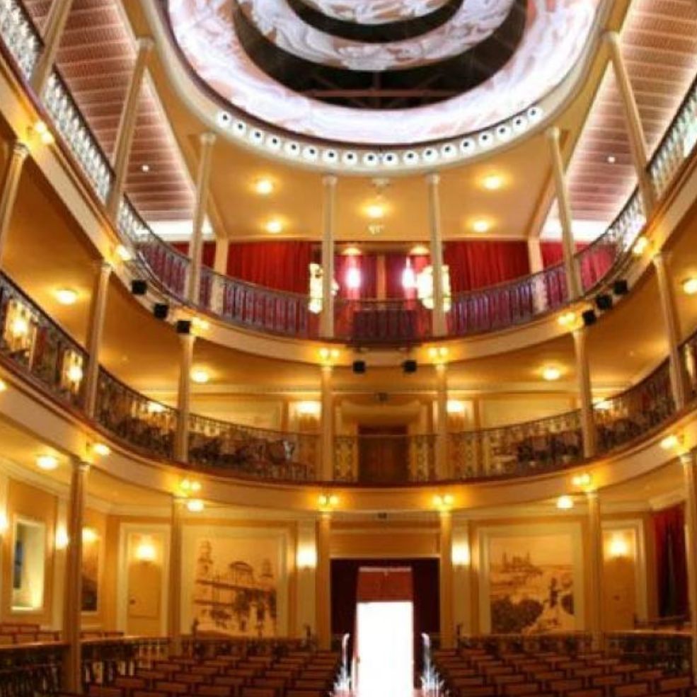 Elegante interior de teatro con balcones ornamentados, luces cálidas y un techo abovedado sobre butacas vacías.