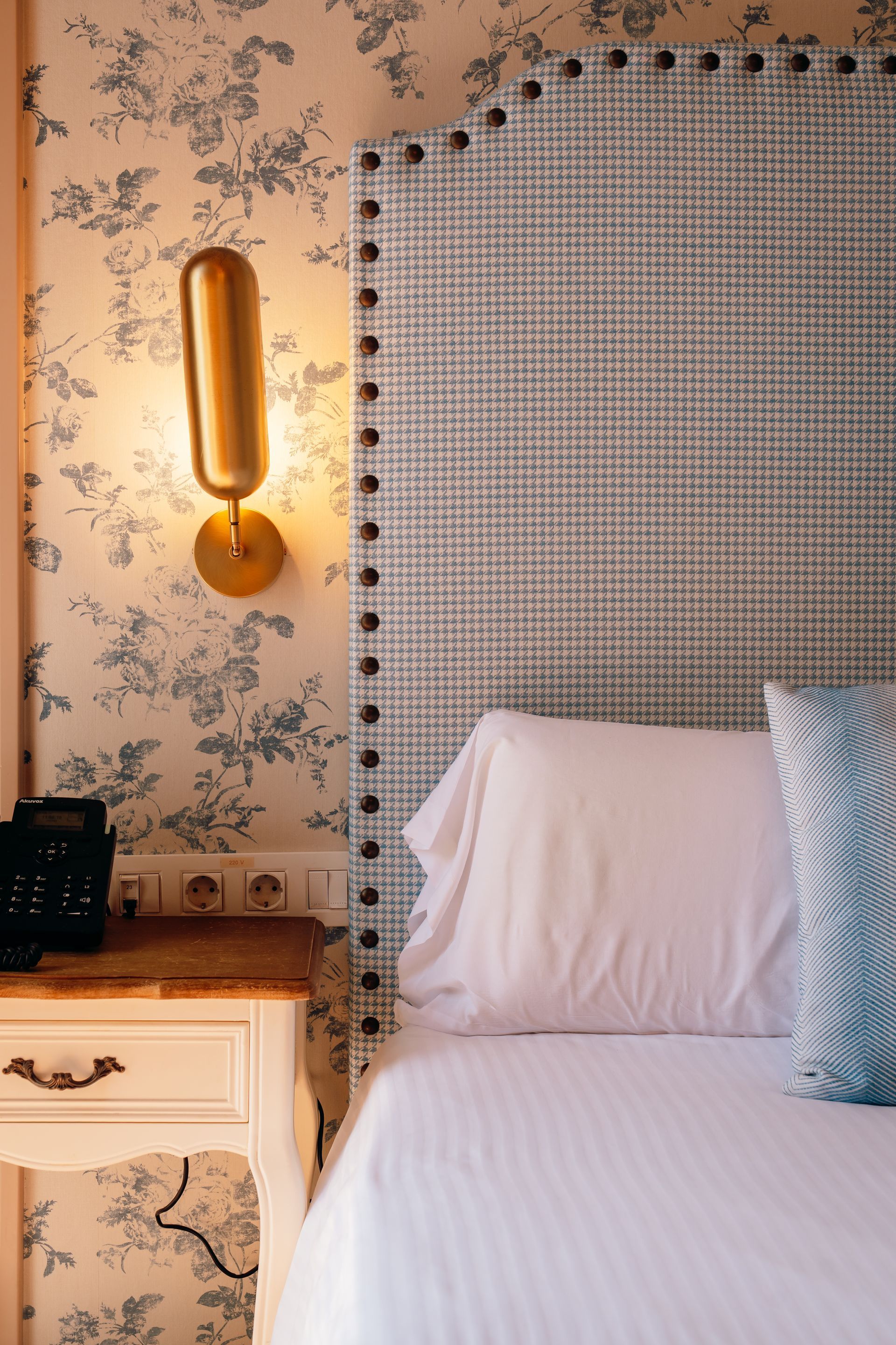 Acogedora habitación con papel pintado floral, aplique de pared iluminado y cama con sábanas blancas.