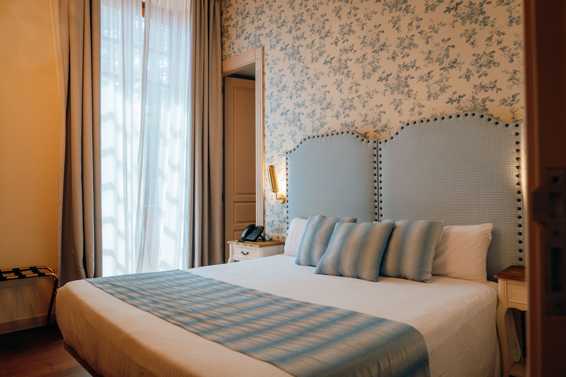 Habitación acogedora con cama, cabecero a rayas, detalles en azul y una gran ventana con cortinas.