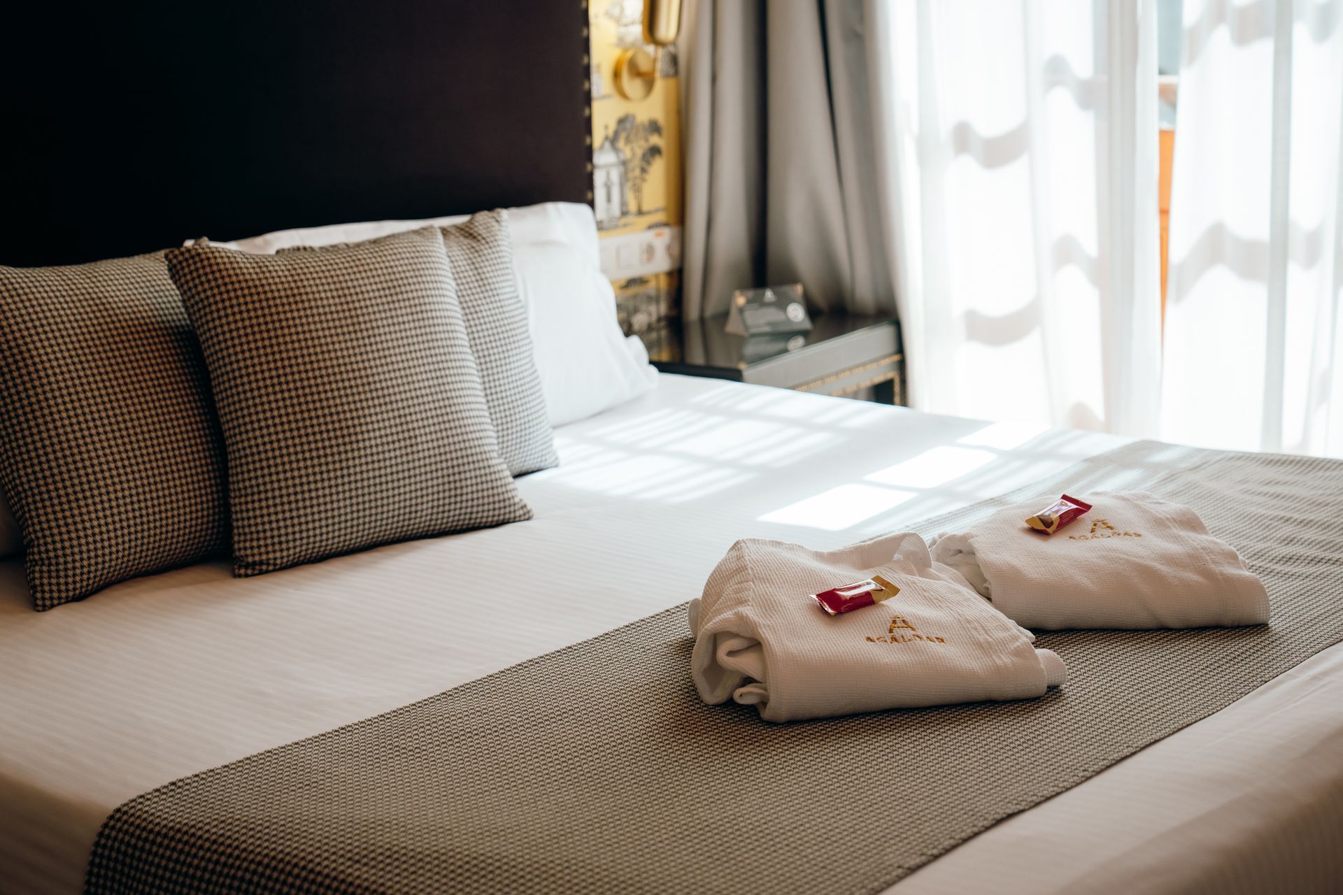 Cama de hotel con sábanas blancas, dos toallas dobladas y luz natural que entra por cortinas transparentes.