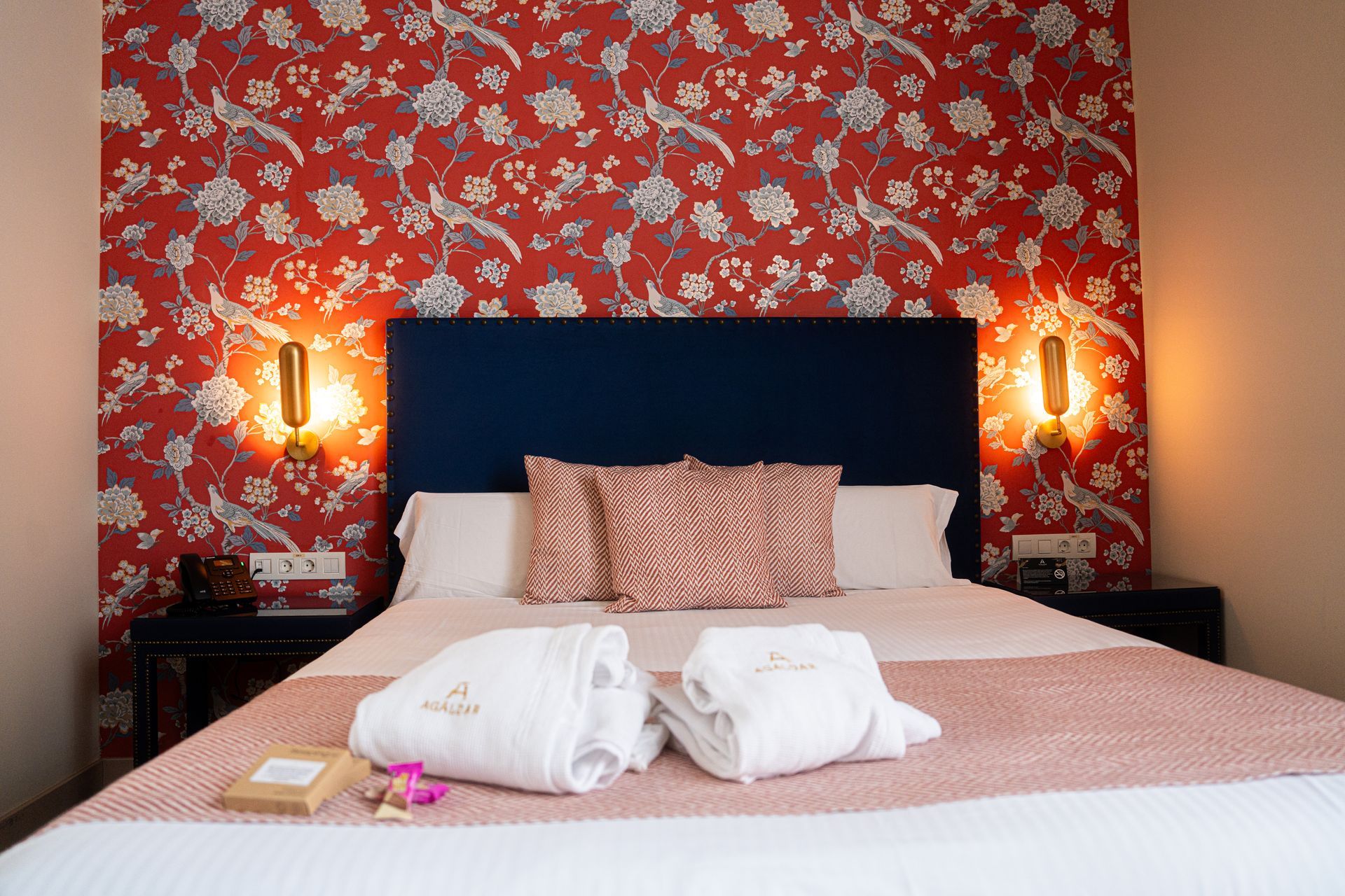 Cama con cabecero azul marino, papel pintado floral rosa y toallas cuidadosamente dispuestas bajo luz cálida.