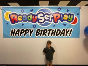 A boy stands in front of a ready set play happy birthday banner