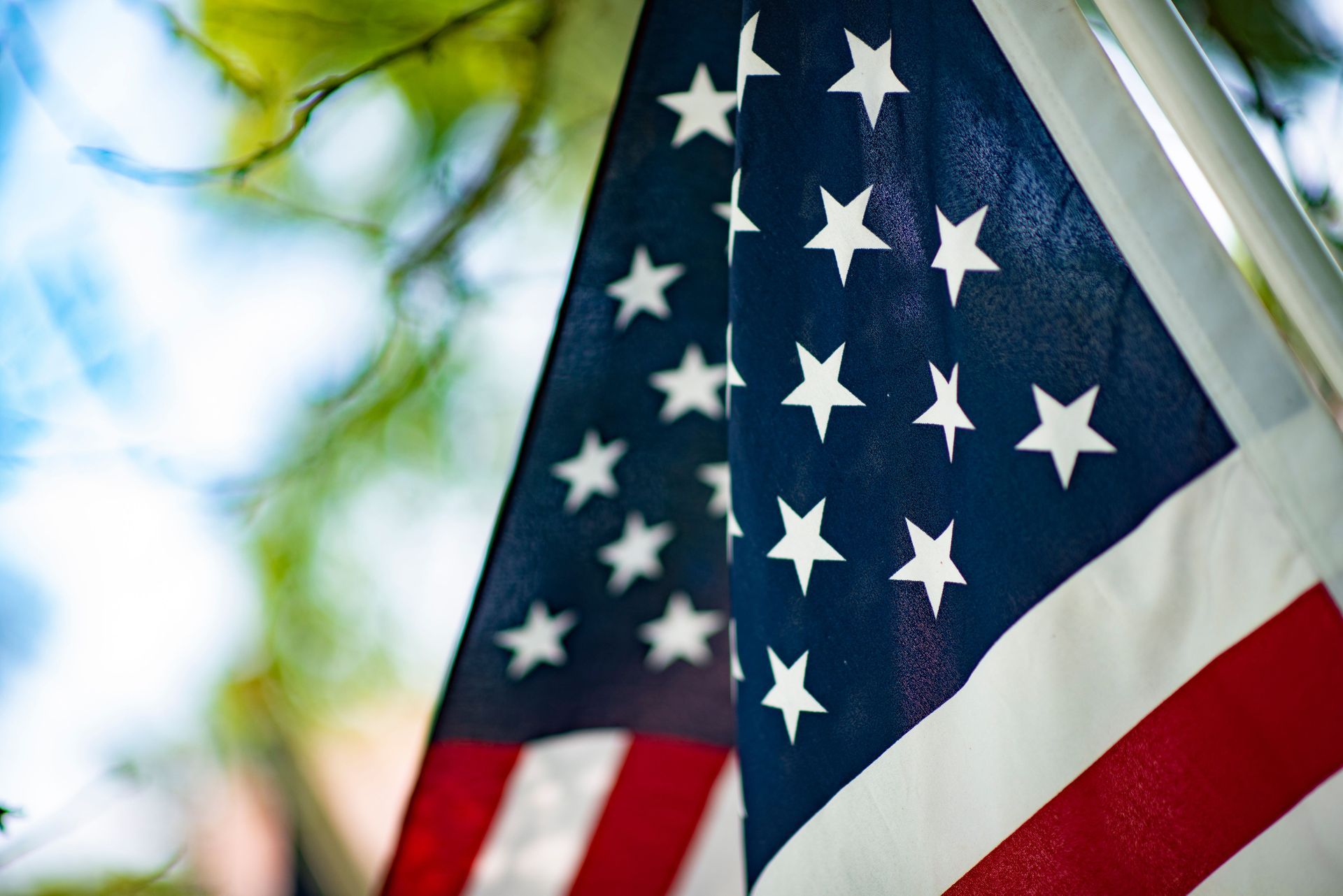 Two American flags waving outdoors with blue sky and blurred trees in the background