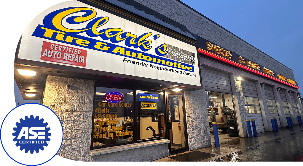 Auto repair shop storefront at dusk with blue awning, bright signage, and ASE logo by the entrance  | Clark's Tire & Automotive