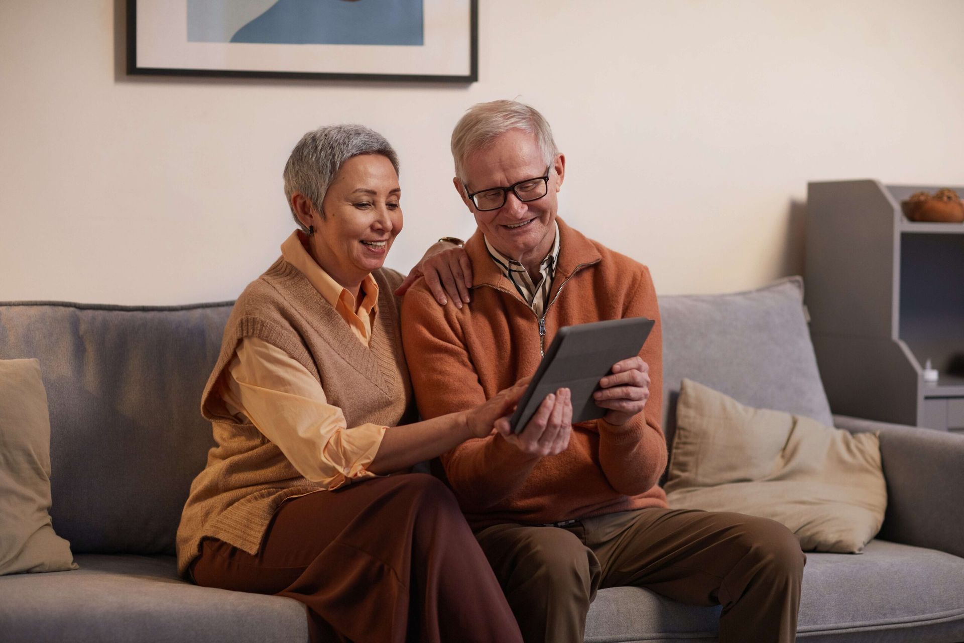 Paar sitzt auf einem Sofa, schaut gemeinsam auf ein Tablet und lächelt. Warm beleuchtetes Wohnzimmer.