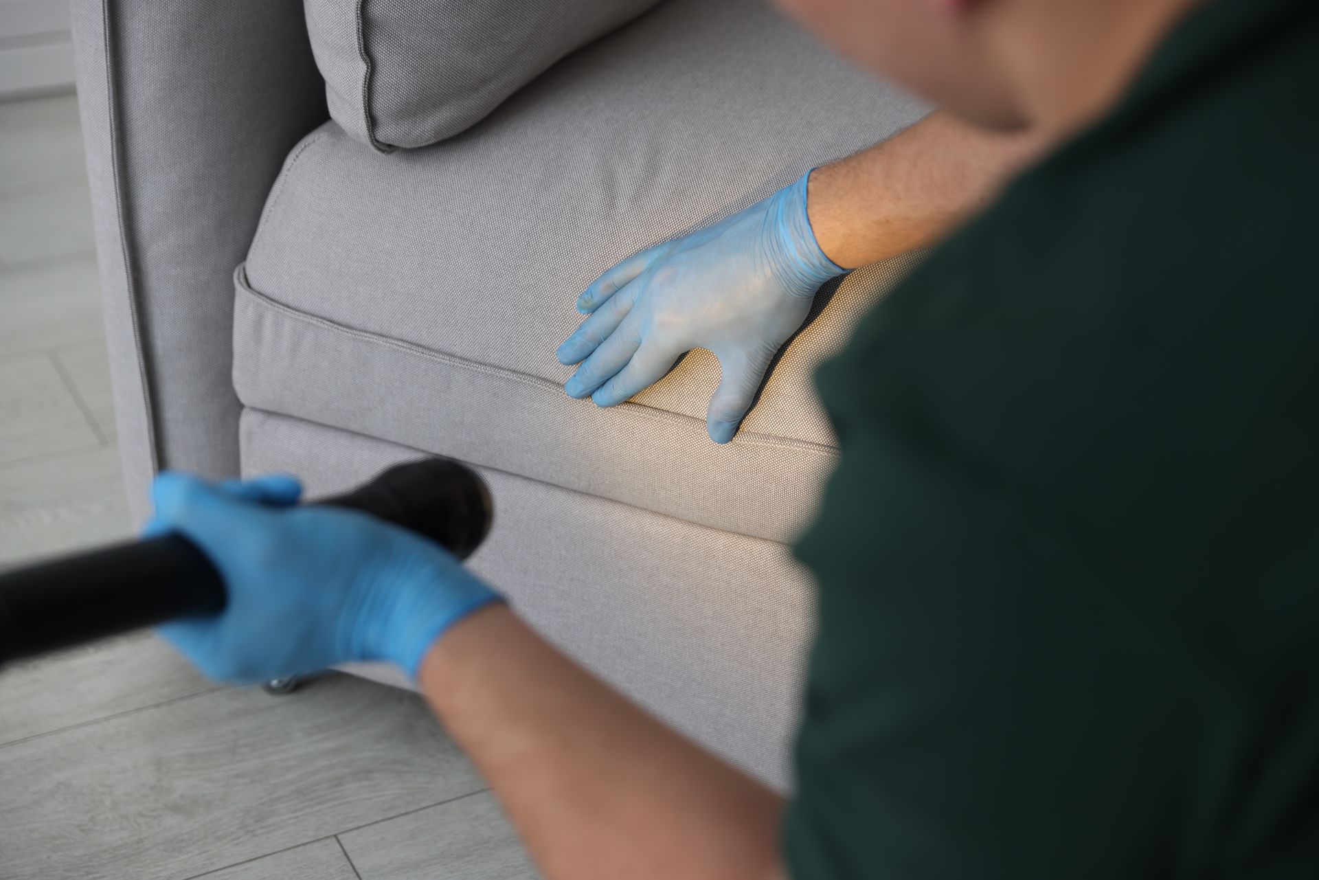 Blue-gloved hands cleaning a beige sofa with a spray bottle and cloth.