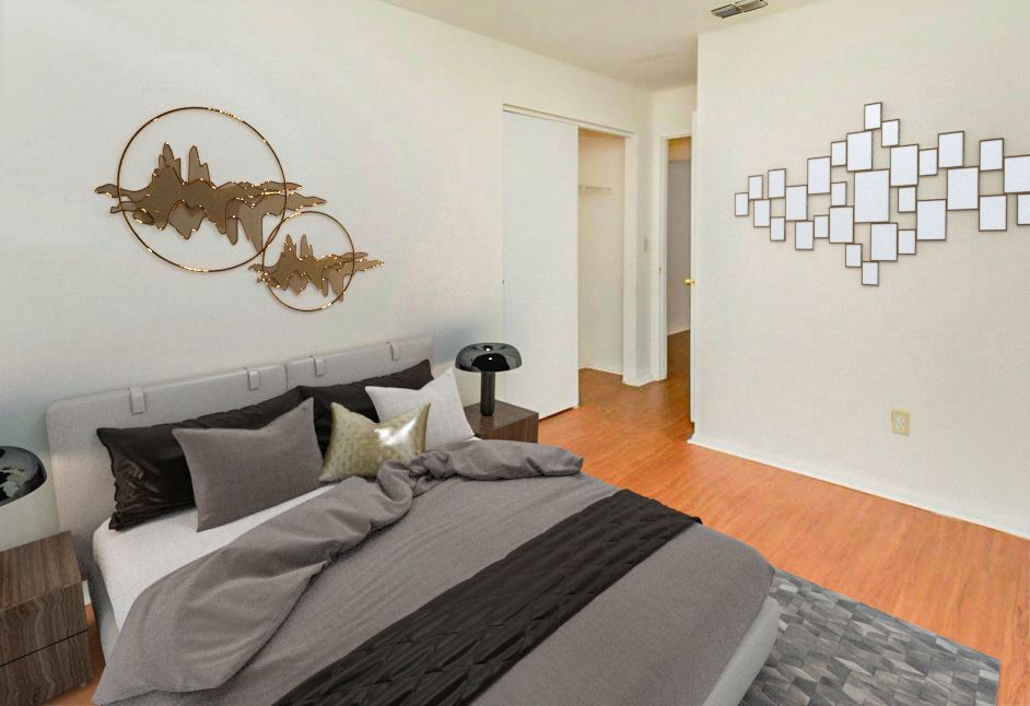 Bedroom with gray bedding, wall art, hardwood floors, and a bedside table with lamp.