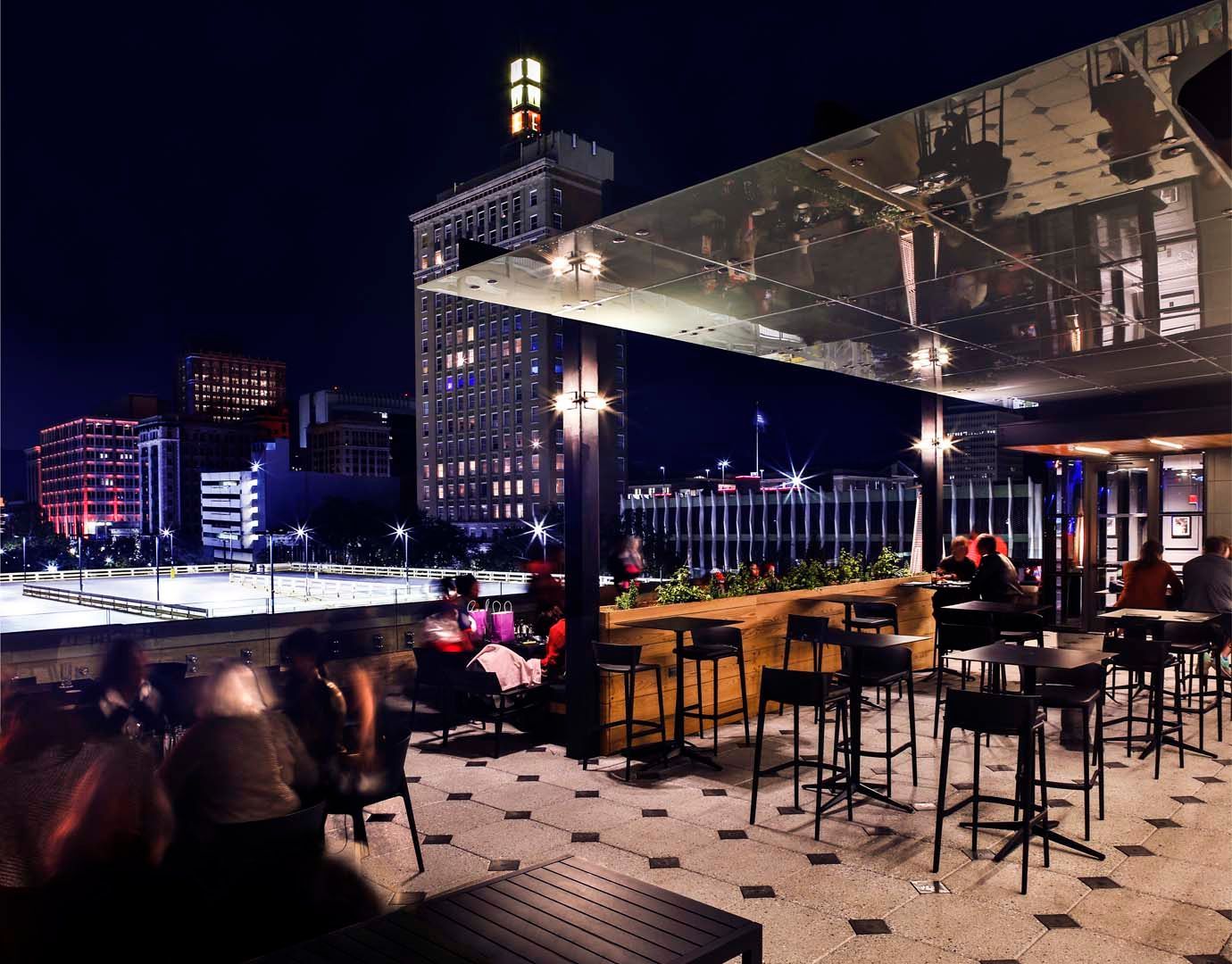 Nighttime rooftop bar with city skyline view; patrons seated at tables and bar stools.