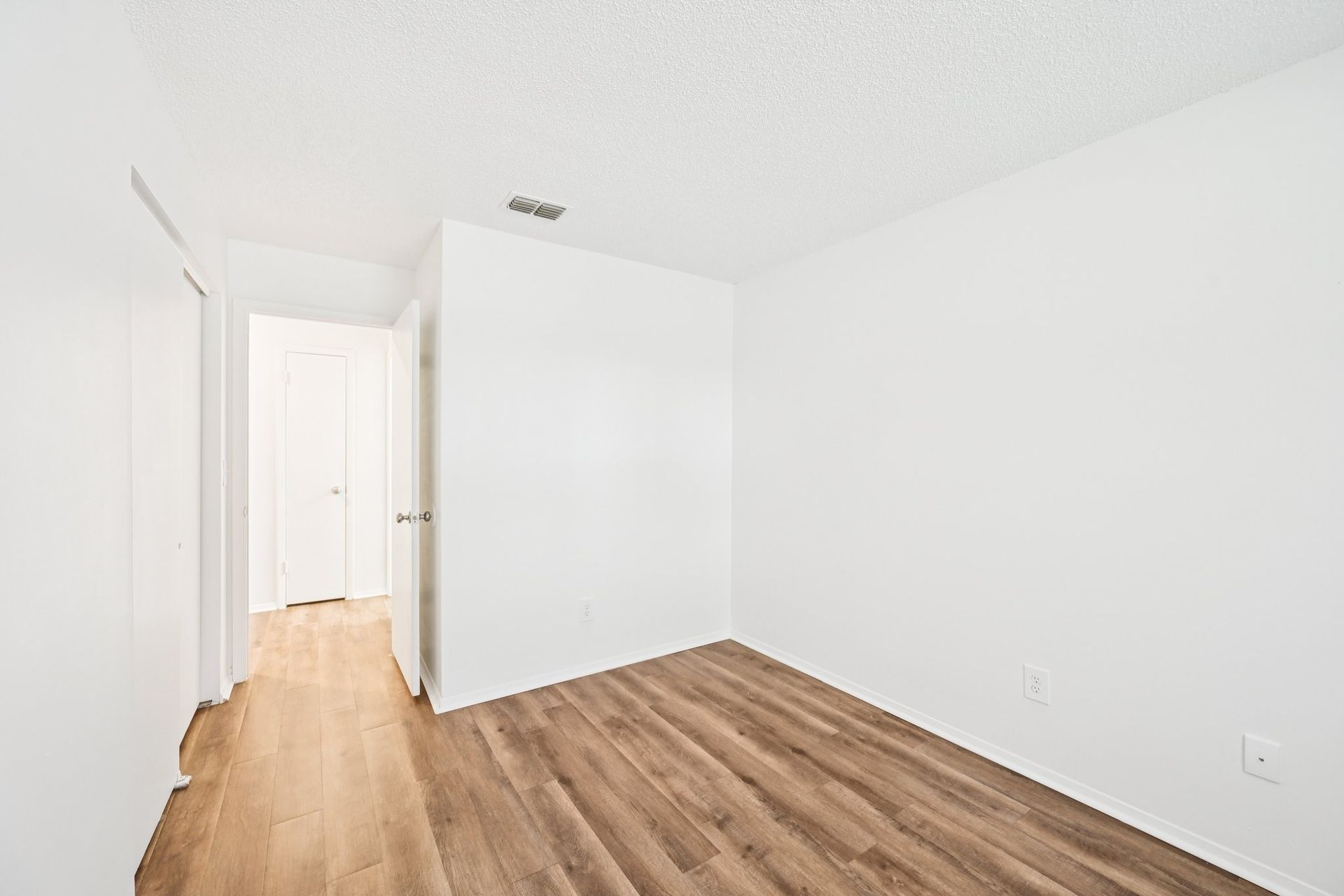 Empty room with wood-look flooring and white walls. A doorway leads to a hallway.