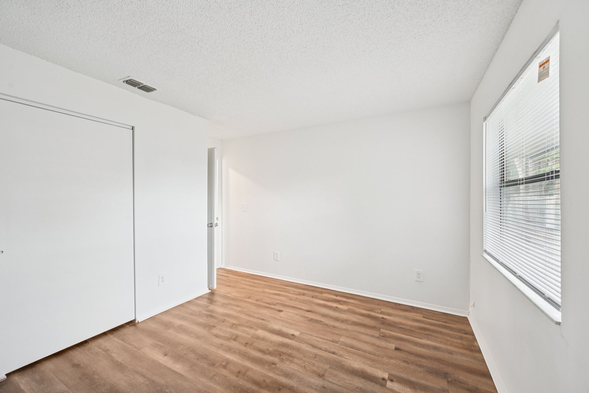 Empty room with white walls, wood-look flooring, closet, and a window with blinds.