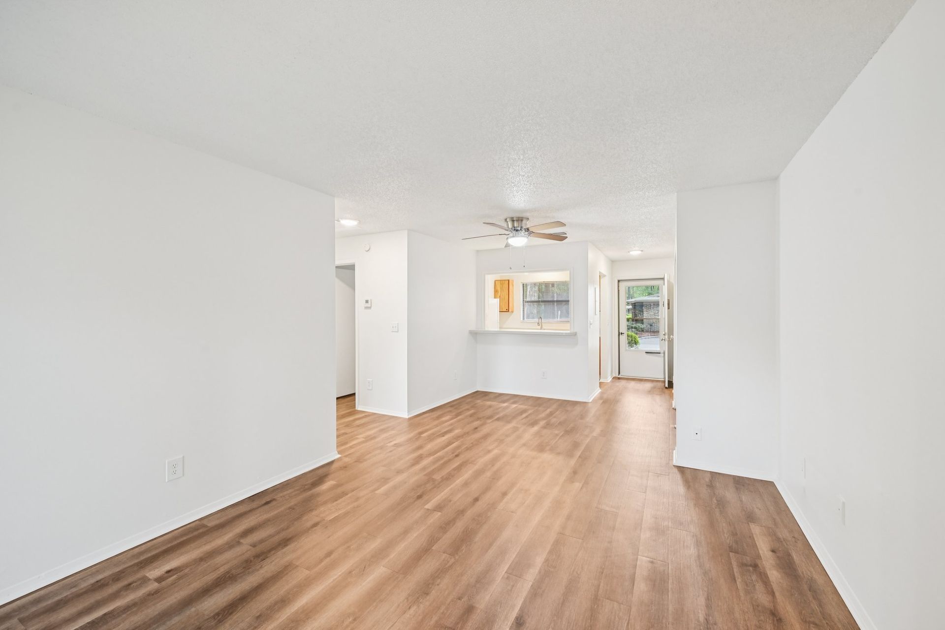 Interior view of an empty apartment with wood-look flooring and white walls, leading to a kitchen and doorway.
