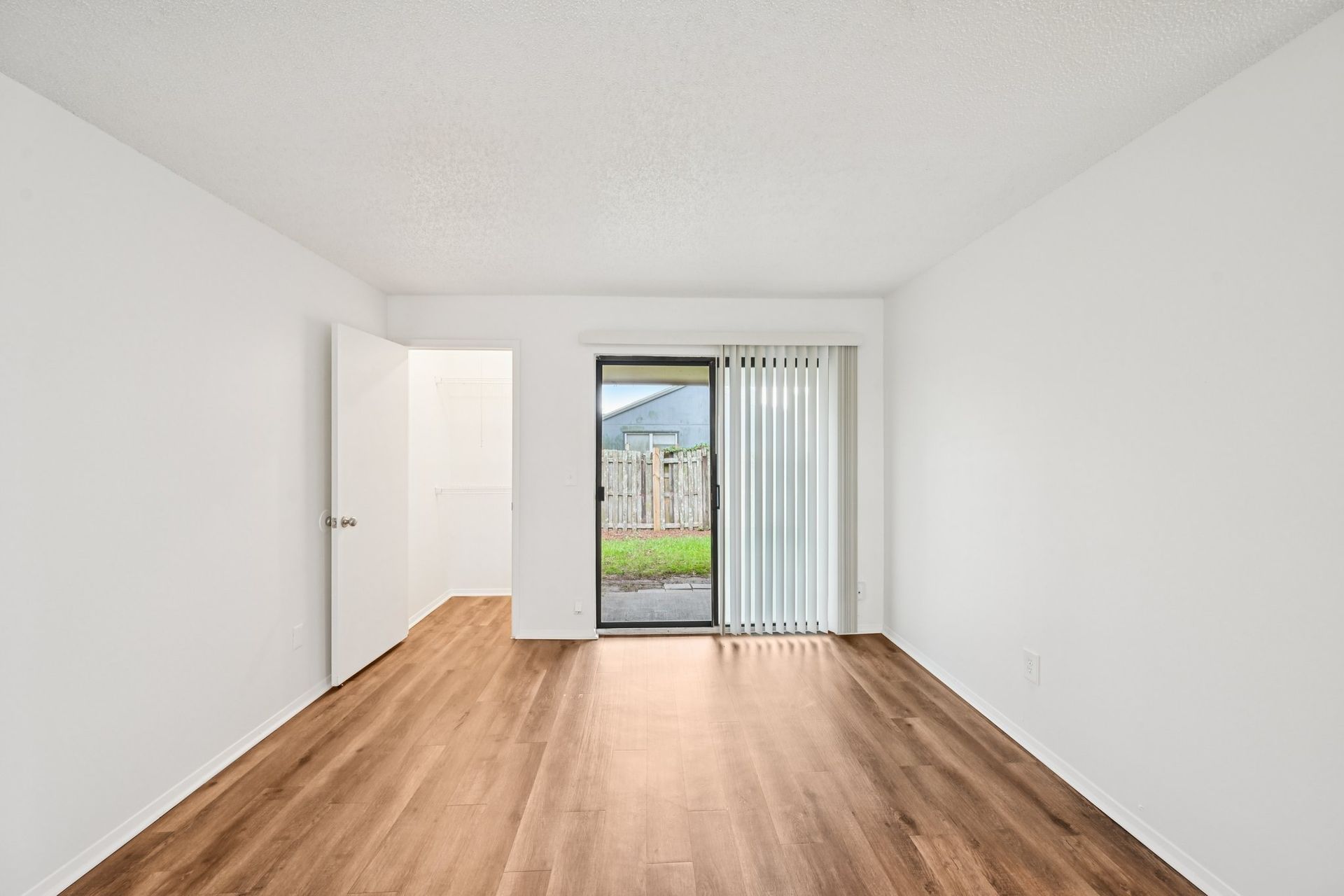 Empty room with wood floors, white walls and ceiling. Sliding glass door to yard, door to interior.