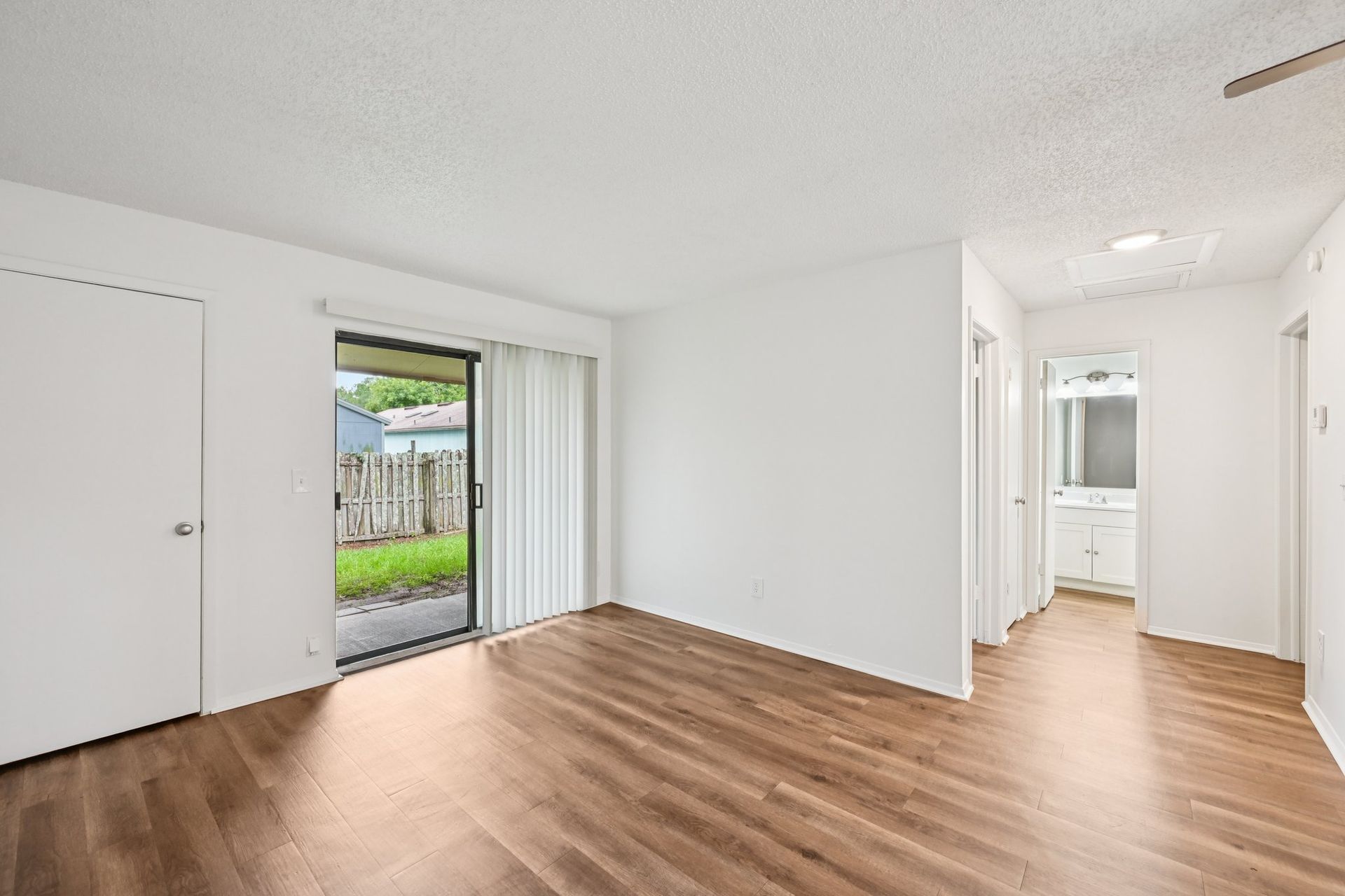 Empty, white-walled room with wood-look flooring and sliding glass door to a small yard.