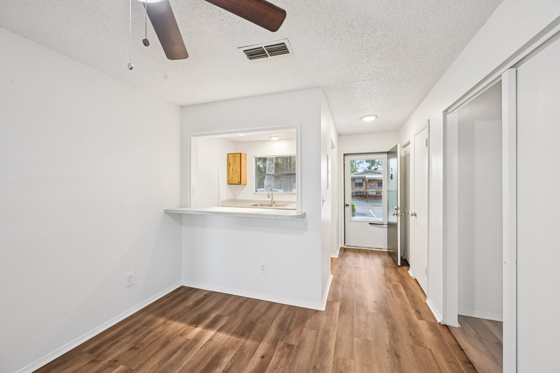 Empty room with wood-look flooring, white walls, and a pass-through to a kitchen.