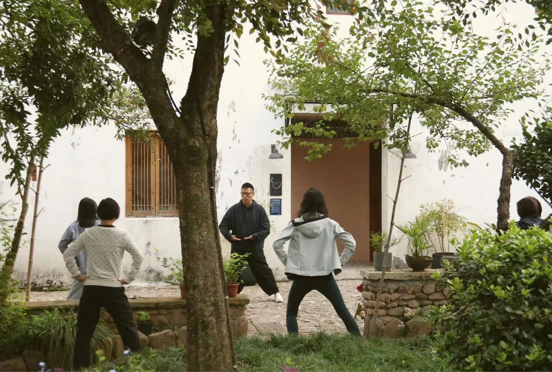 People standing in a yard facing a man by a white building with trees and stone steps.
