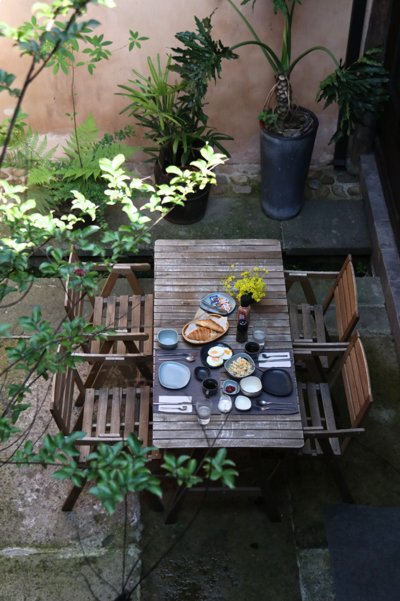 Outdoor patio with a wooden table set for tea, surrounded by potted plants and chairs