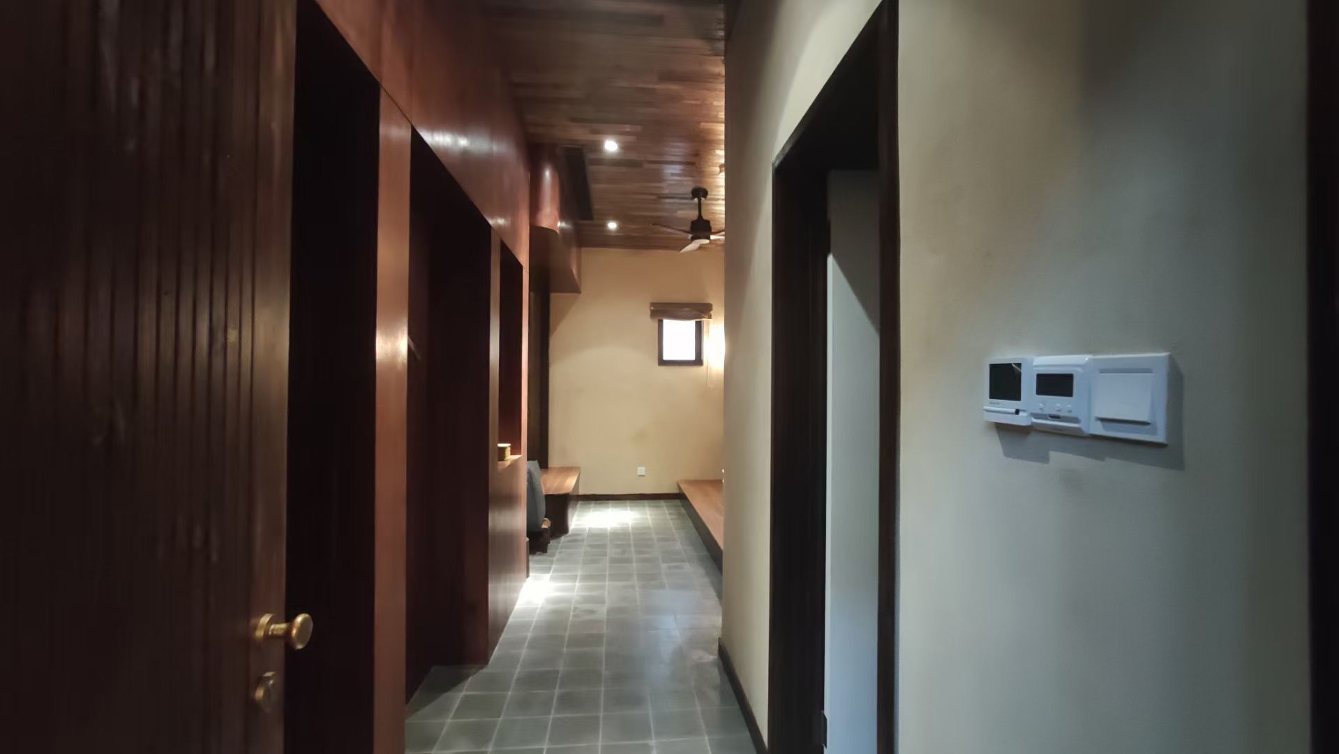 Dim hallway with wood-paneled doors, tiled floor, and wall-mounted controls in a modern interior.