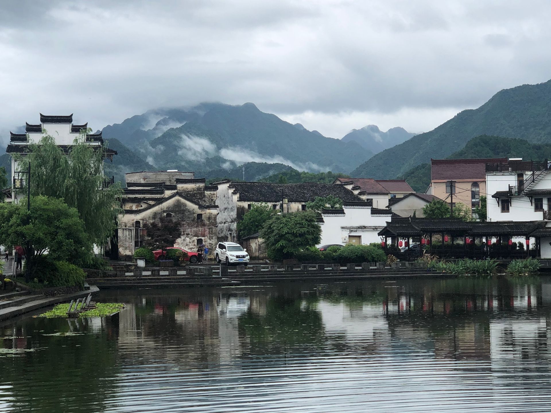 Lakeside village with buildings and mountains reflected in calm water under cloudy skies
