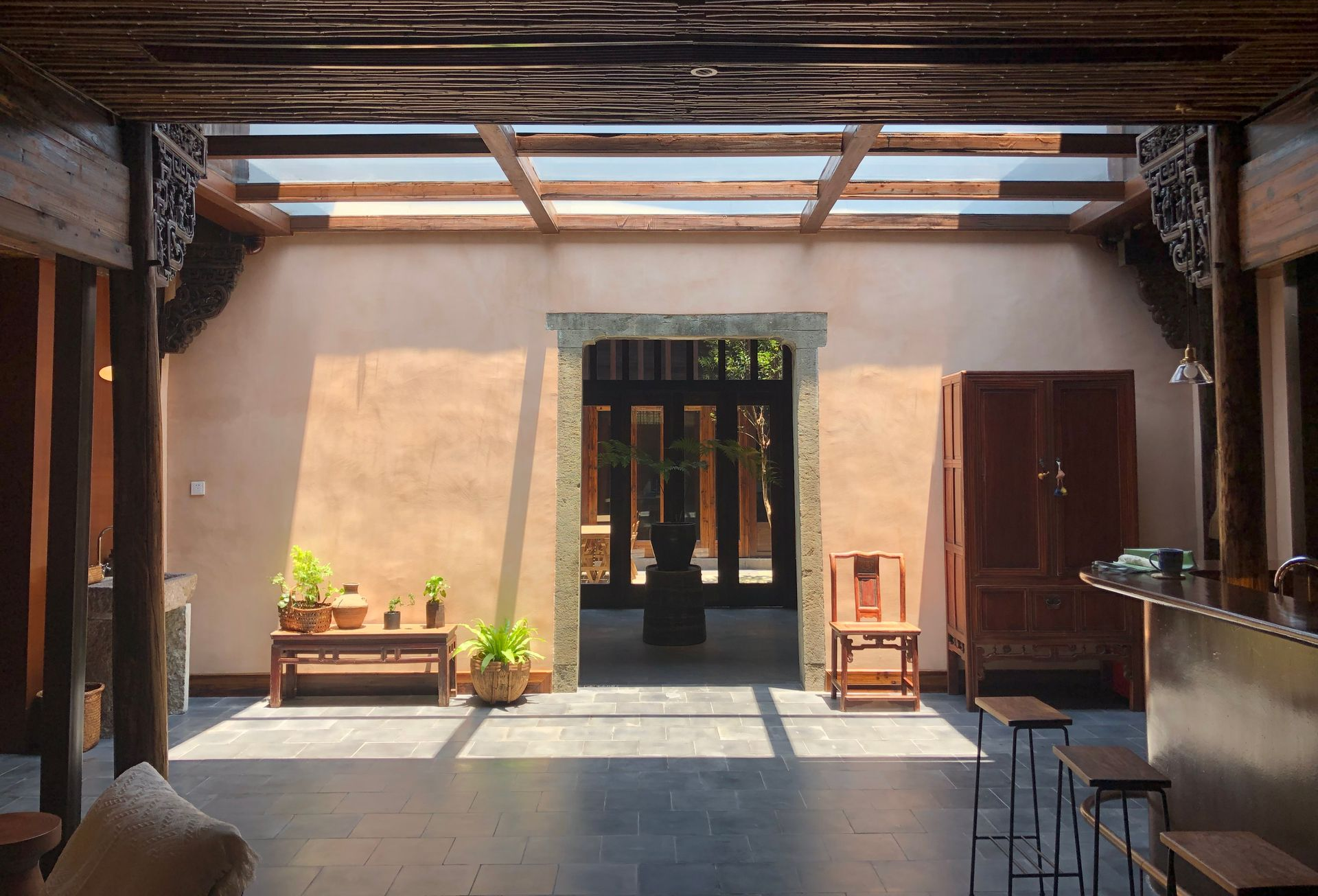 Sunny courtyard with wooden beams, potted plants, and open doorways leading into a rustic home.