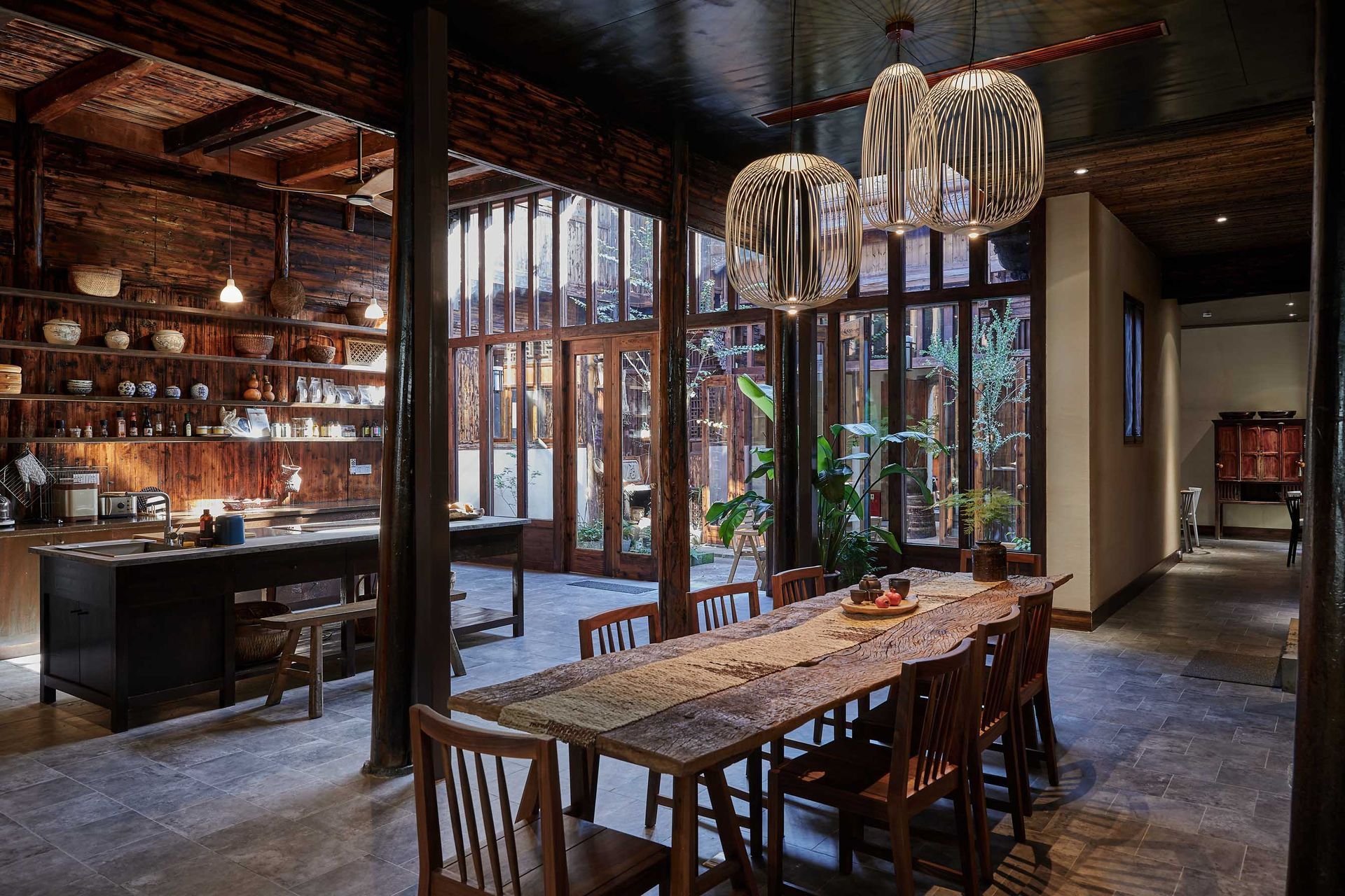 Warm, upscale dining room with wooden tables, candles, hanging lights, and large windows overlooking greenery