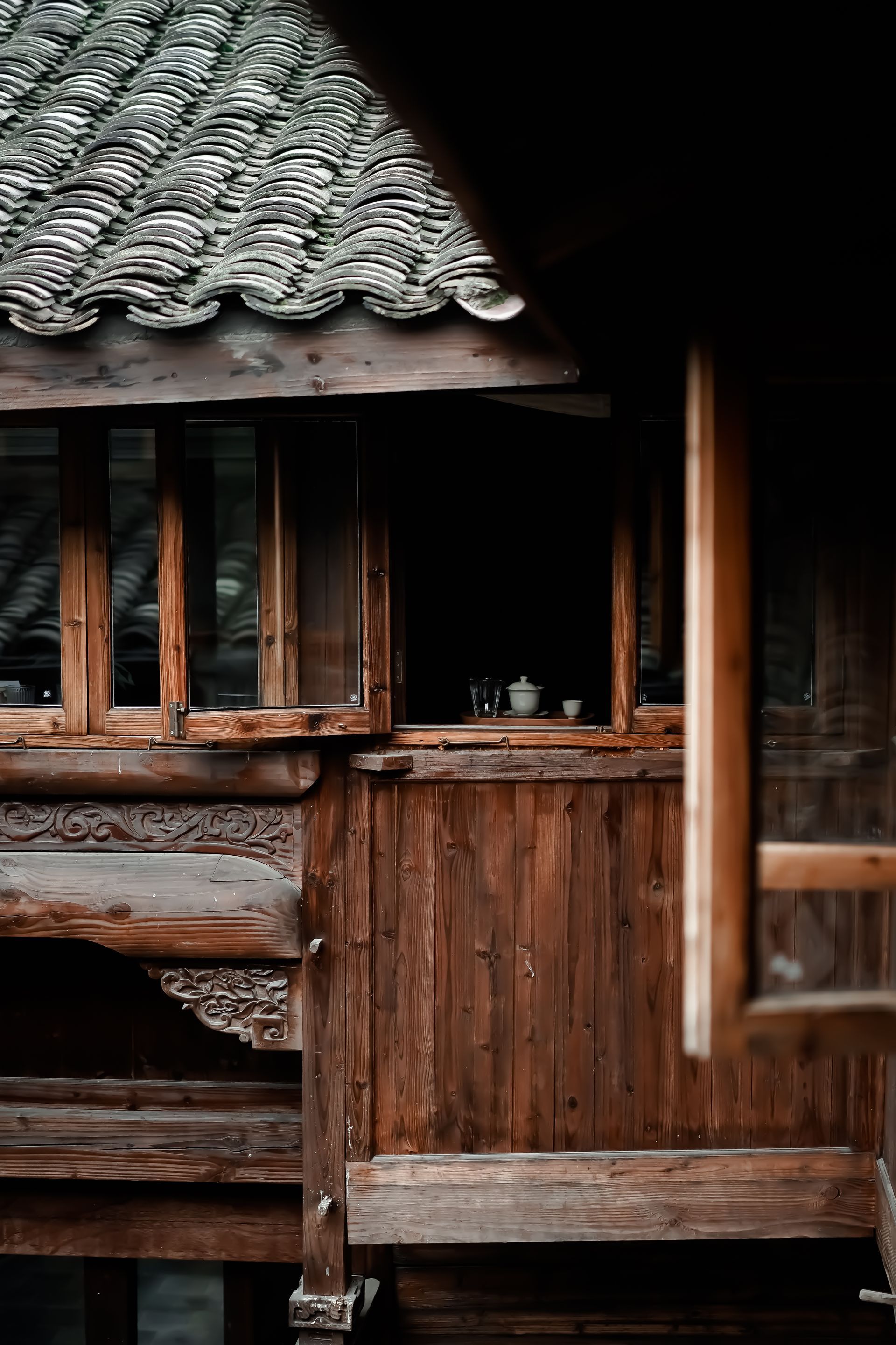 Rustic wooden cabin porch with small window and weathered shingled roof, partly in shadow