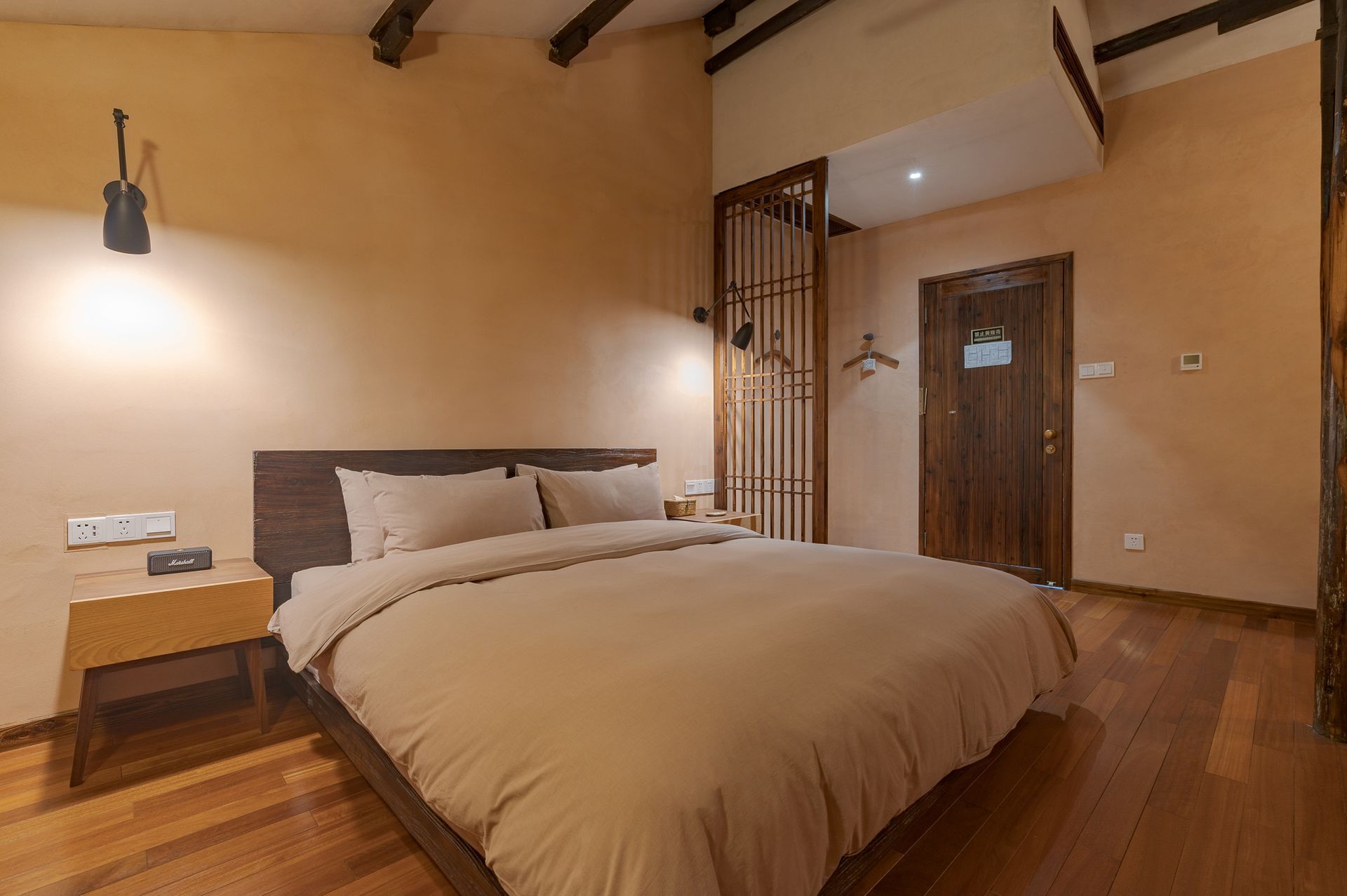 Cozy hotel bedroom with a large bed, warm wood floors, beige walls, and a wall-mounted lamp