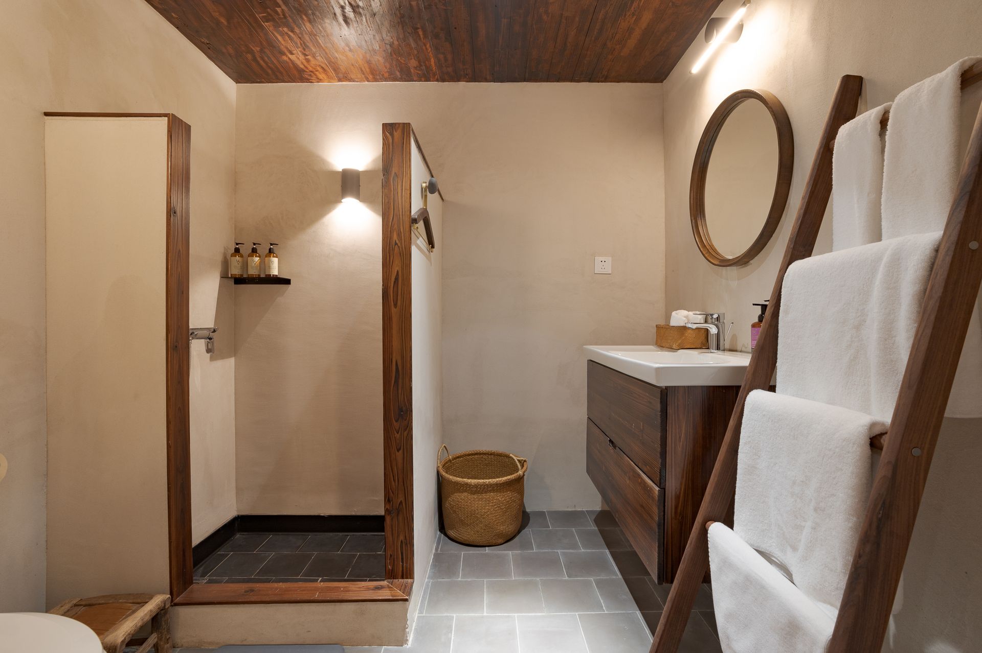 Modern bathroom with wood vanity, round mirror, shower, and white towels on a ladder rack.
