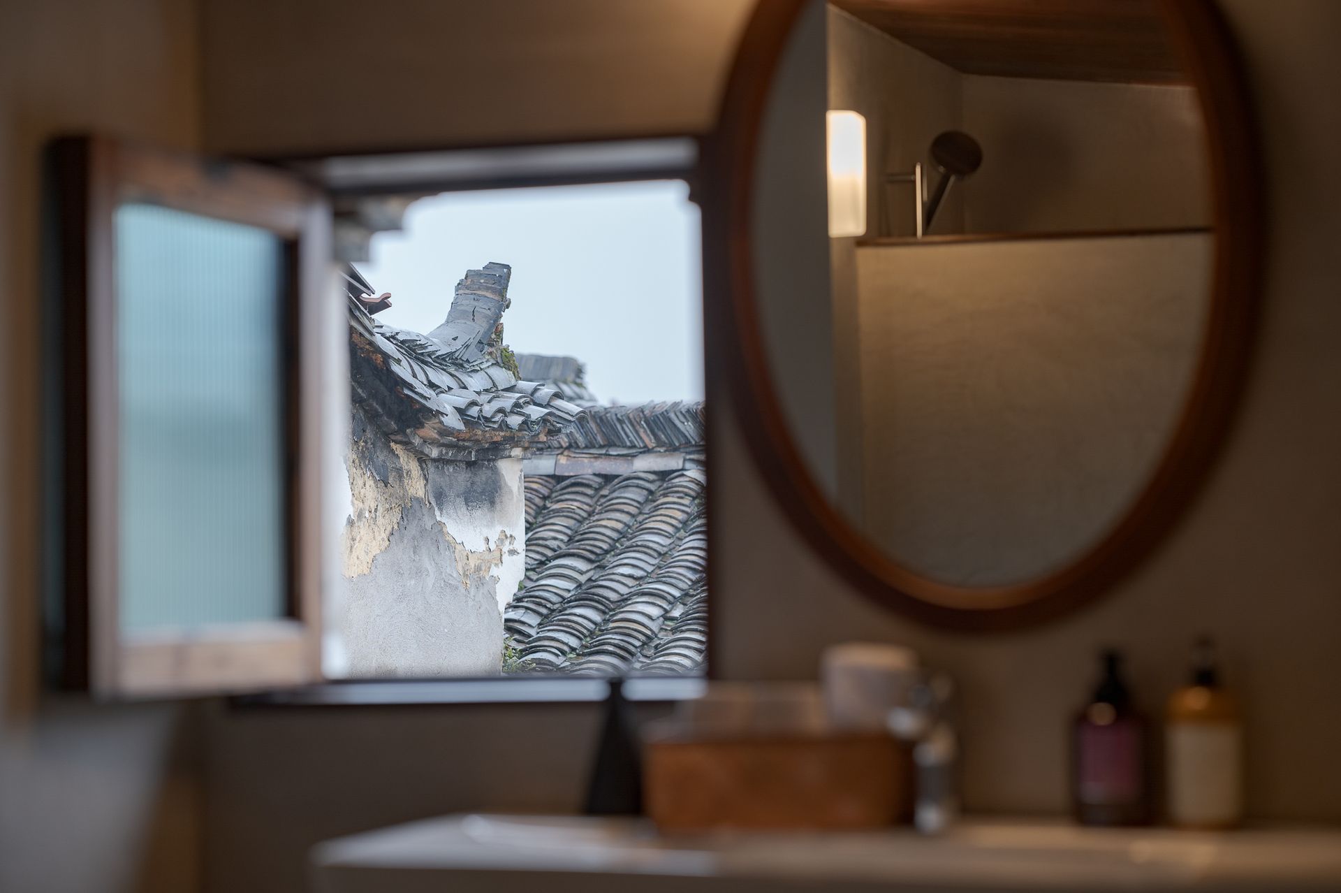 Window view of snowy rooftops and trees, seen from a bathroom with a round mirror and sink.