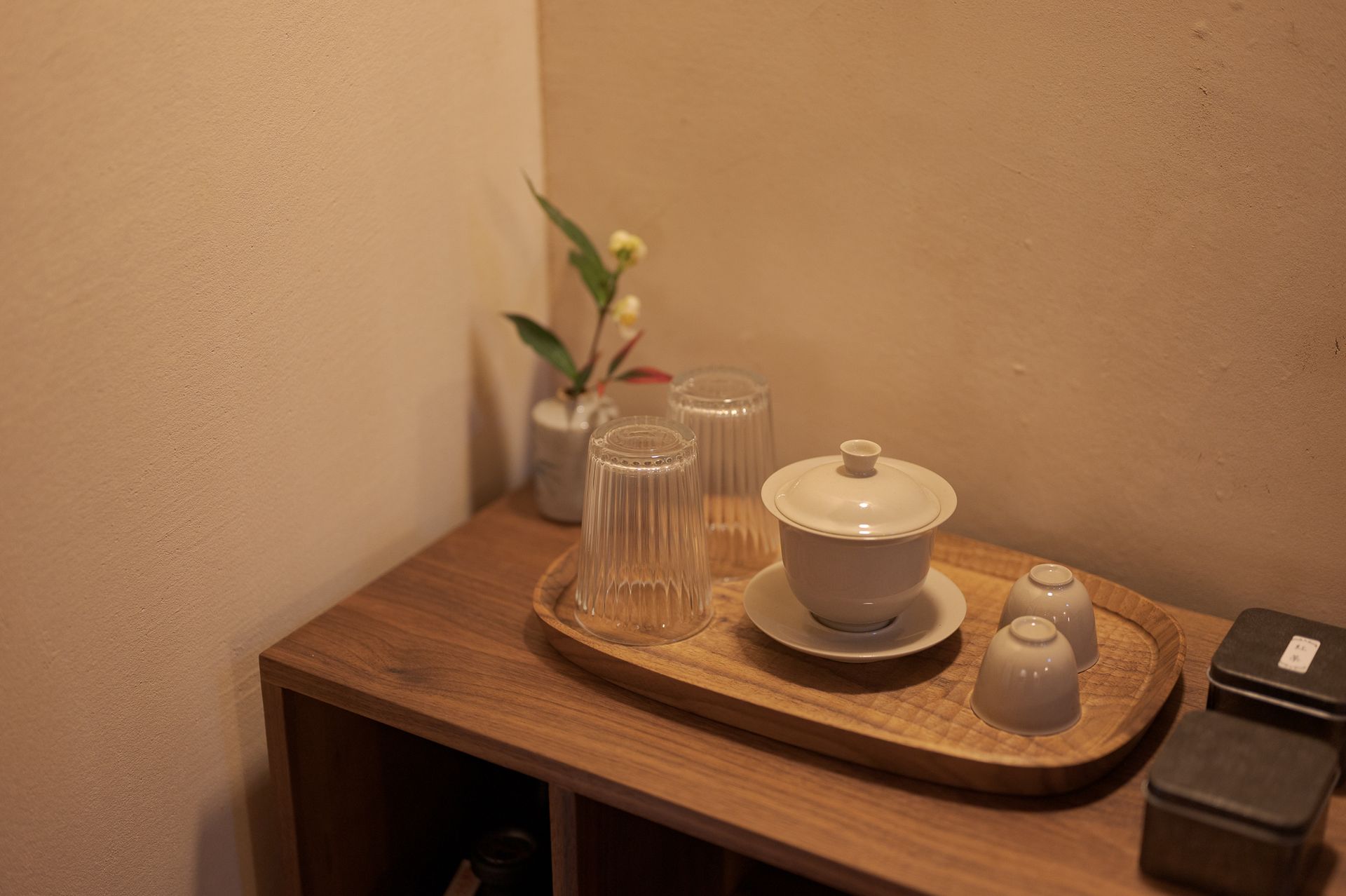 Tea set on a wooden tray beside a small vase on a shelf in a warm-toned room