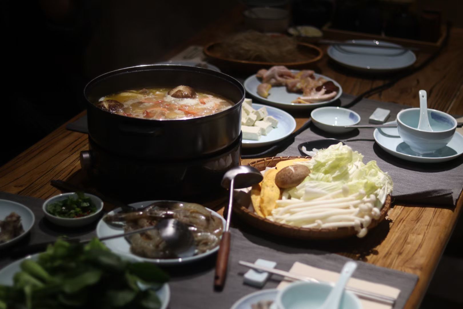 Hot pot meal on a wooden table with vegetables, meat, dipping sauces, and bowls around it