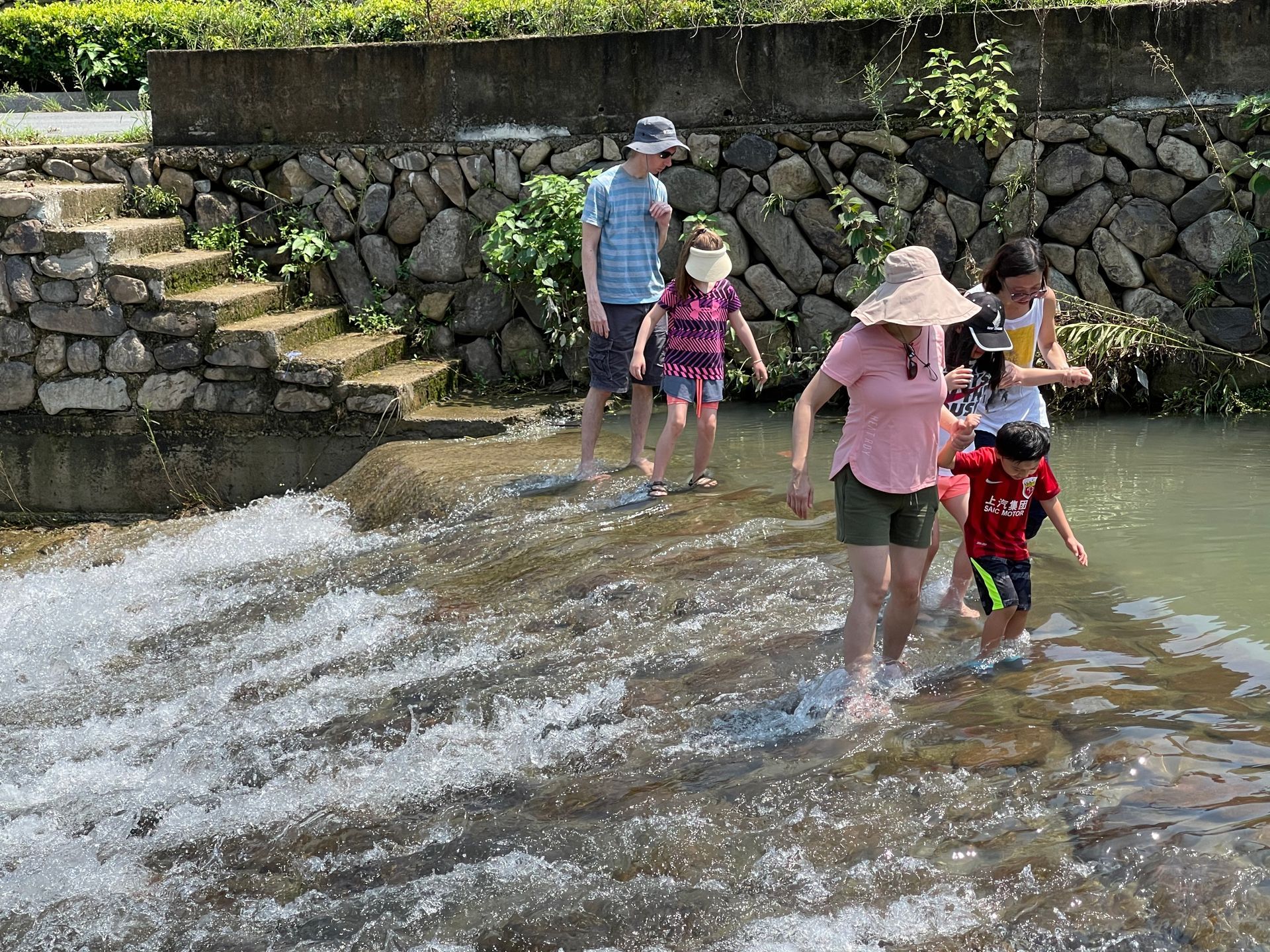 People wading in a shallow stream beside a stone wall and steps on a sunny day