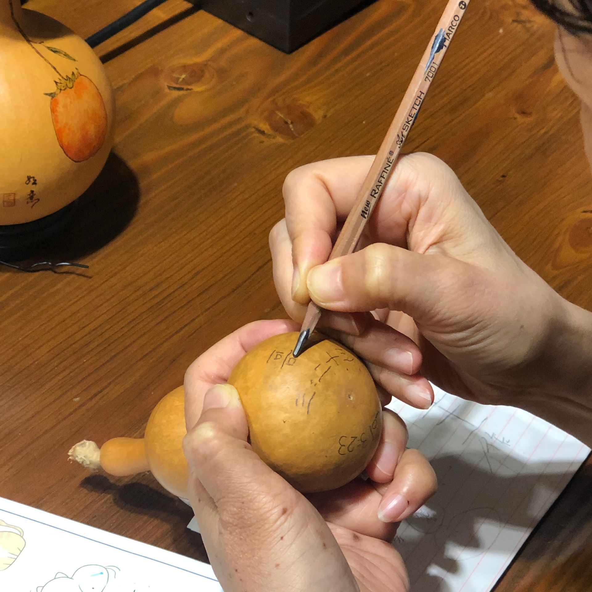 Hands carving a design into a tan fruit on a wooden table, with another fruit nearby.