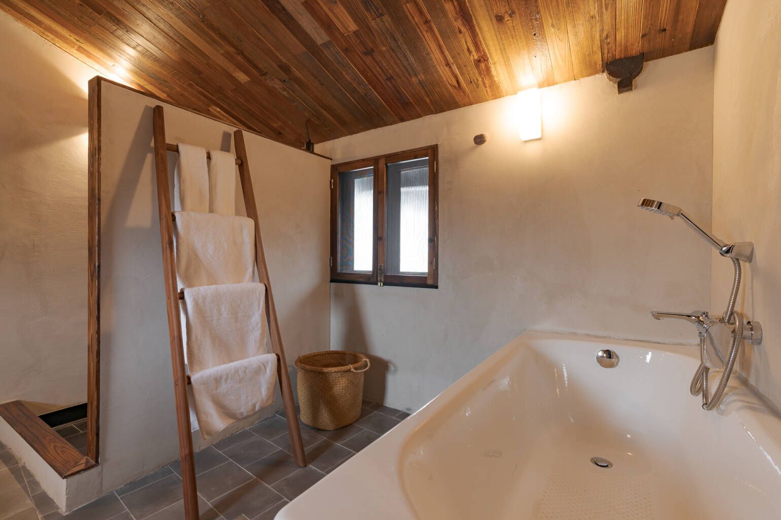 Small bathroom with a bathtub, window, ladder shelf, and sloped wood ceiling.