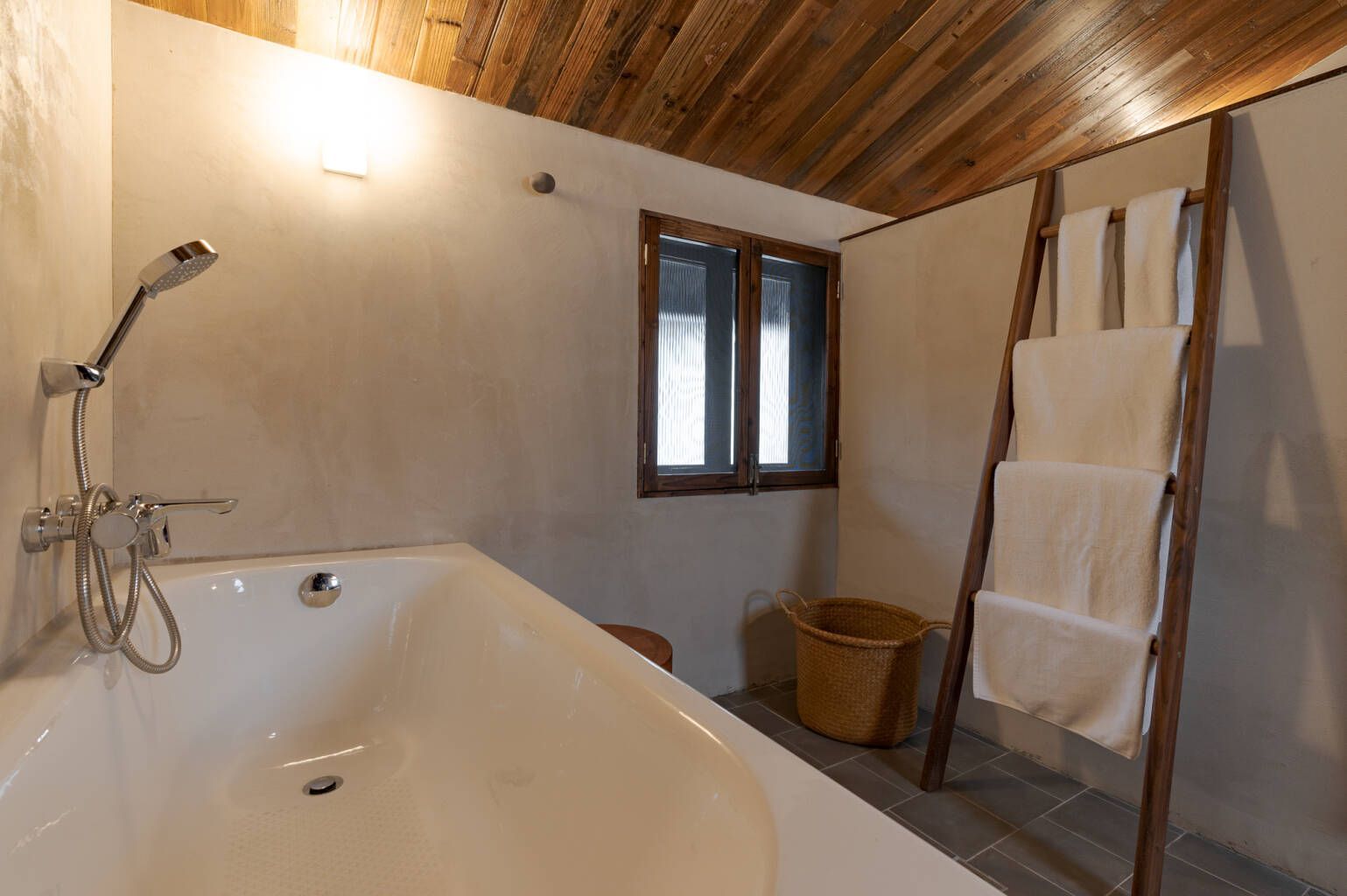 Rustic bathroom with bathtub, showerhead, window, wicker basket, and ladder-style towel rack