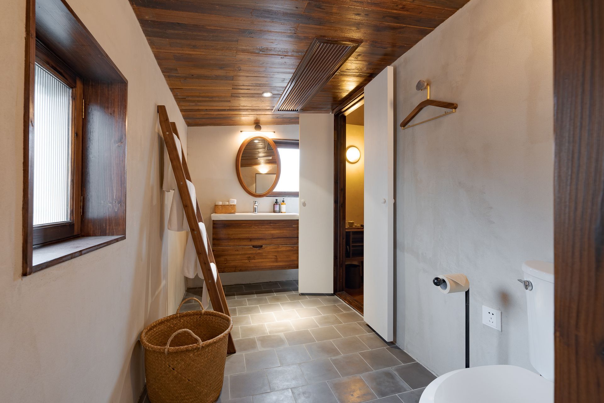 Narrow bathroom with wood ceiling, tiled floor, sink, round mirror, ladder, and wicker basket.