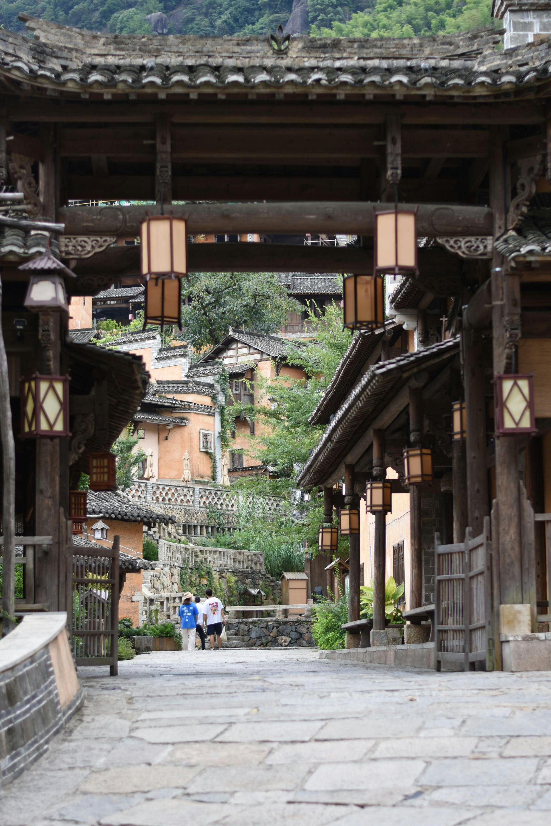 Stone path through a traditional village gate, with old wooden buildings and distant people below green hills.