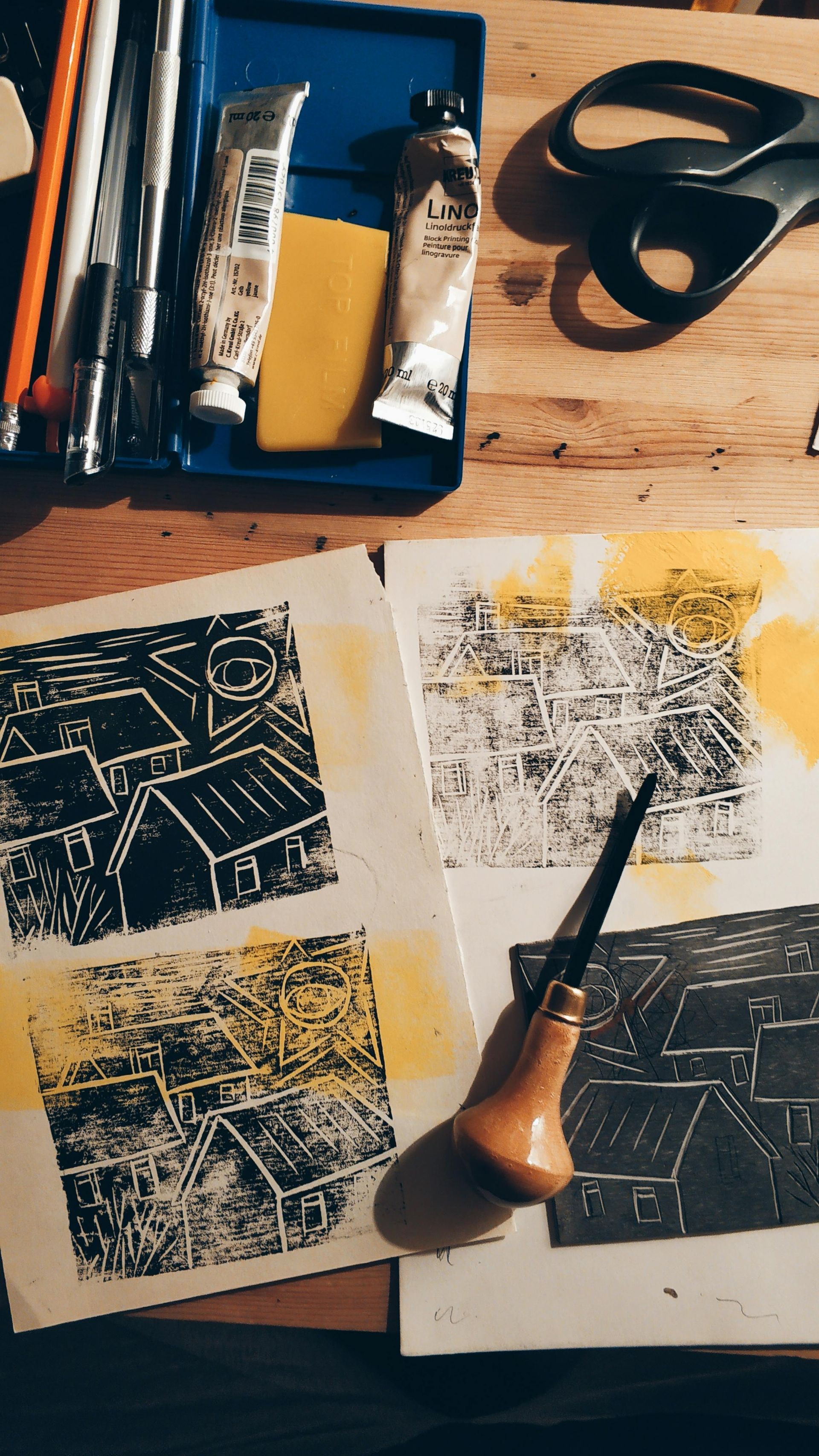 Printmaking tools and carved blocks on a wooden table beside two black-and-white prints