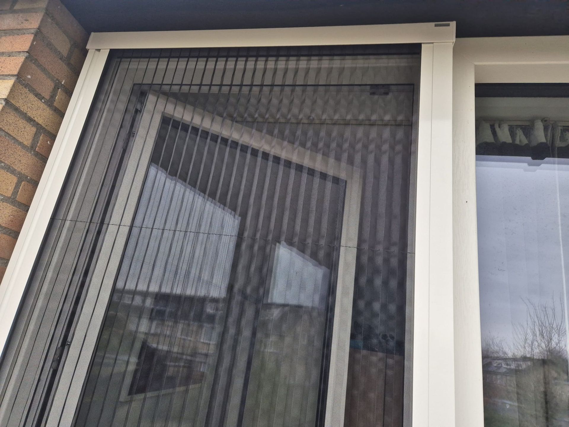 Een raam met horgaas en witte kozijnen naast een bakstenen muur, met een weerspiegeling van de buitenlucht.