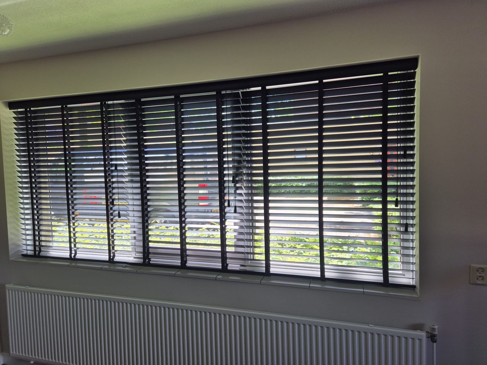 Interieur met een breed raam, zwarte jaloezieën en een witte radiator eronder.