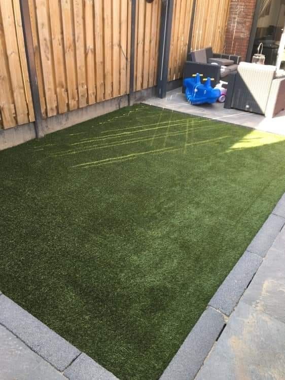 Een kleine hoek met kunstgras, een houten schutting, grijze bestrating en blauwe tuinspullen achterin.