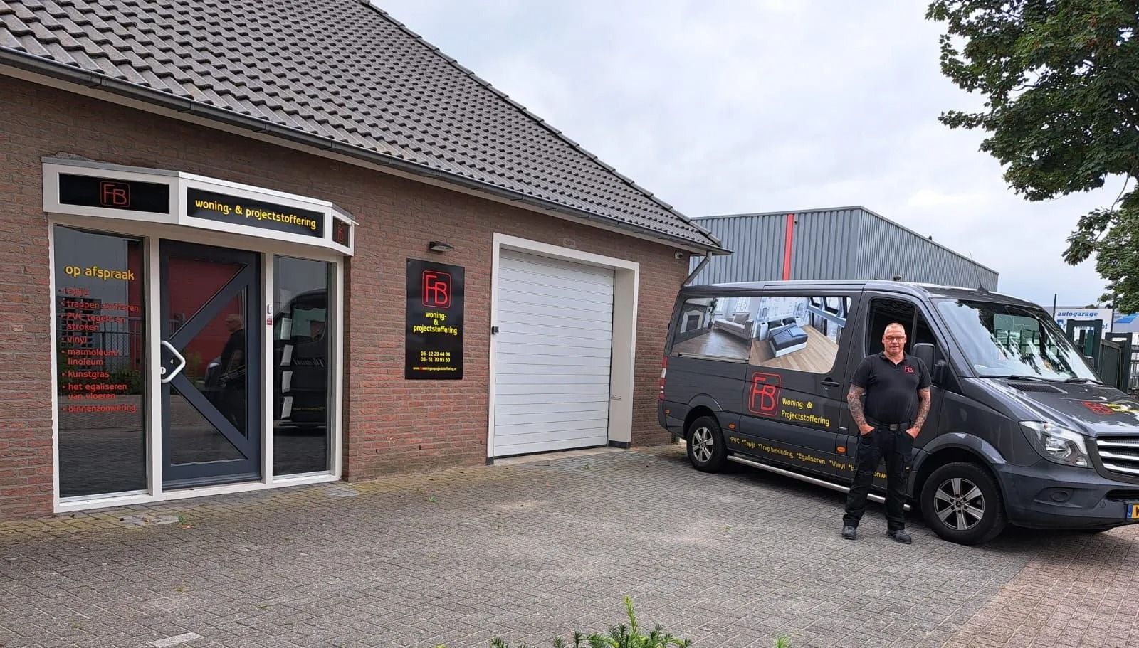Een persoon staat naast een zwarte bestelwagen voor een bakstenen winkel met een garagedeur.