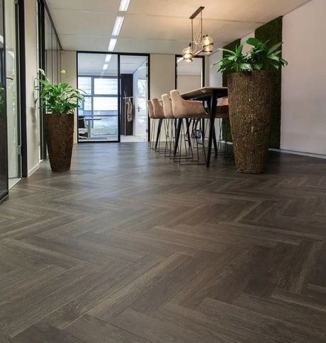 Moderne kantoorlounge met visgraatparketvloer, hoge planten, barkrukken en hanglampen.