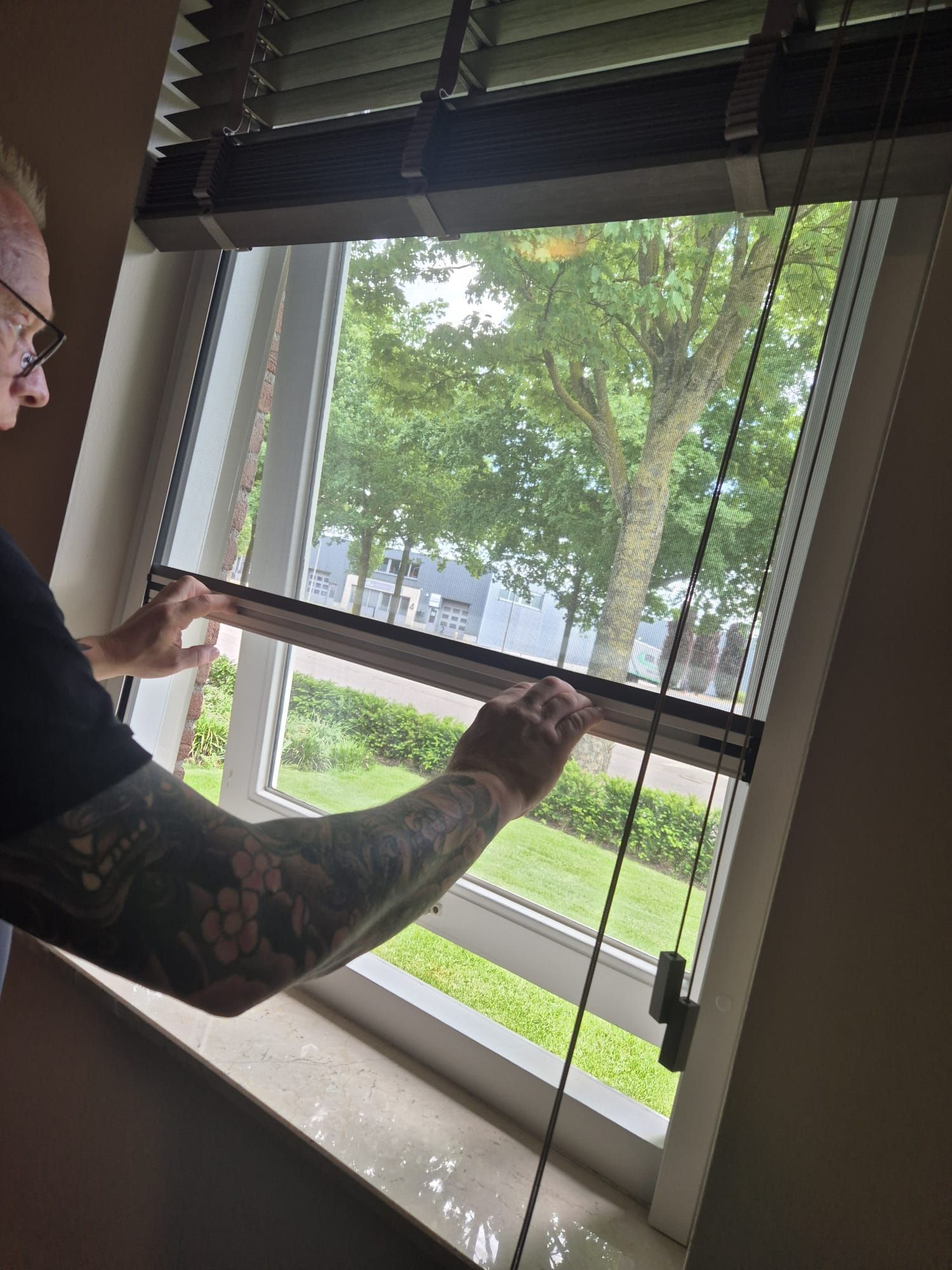 Iemand die een rolgordijn naast een open raam verstelt, met bomen buiten zichtbaar.